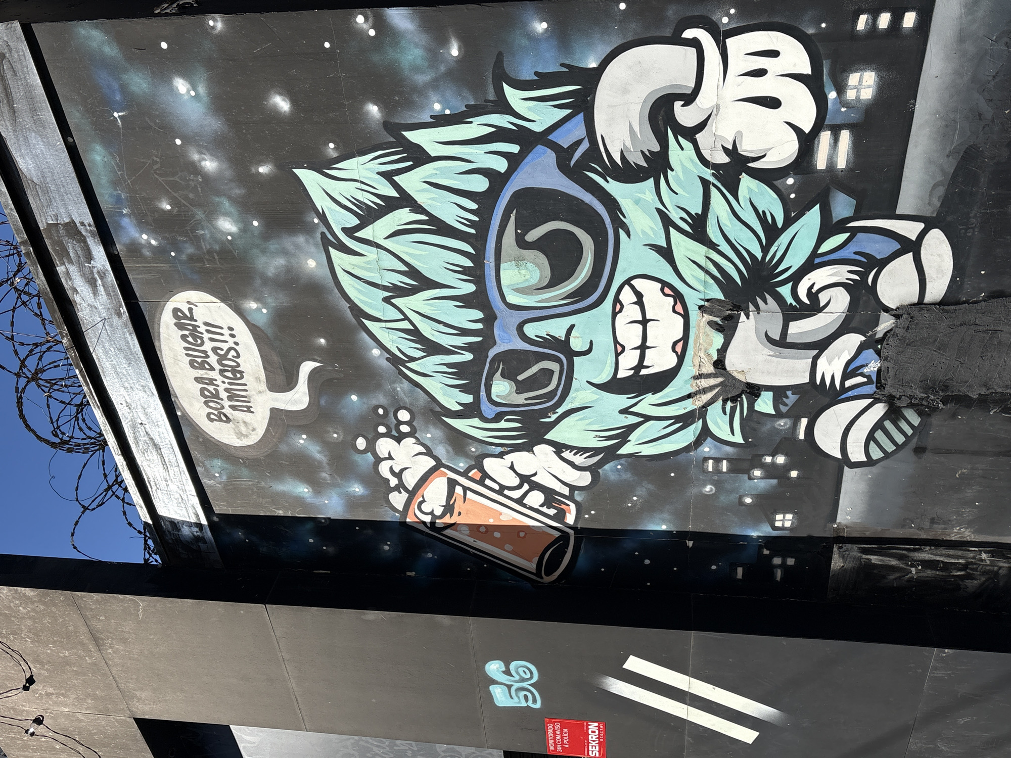 The image shows a graffiti artwork on a black wall in São Paulo, Brazil. The artwork depicts a cartoonish, light blue, plant-like character with large sunglasses, a wide smile, and mint green hair. The character is holding a glass of orange liquid, possibly a drink, in its right hand and appears to be giving a thumbs-up with its left hand. Above the character's head, a speech bubble contains the Portuguese phrase "BORA BUGAR, AMIGOS!!!", which translates to "Let's bug out, friends!!!" The background of the artwork is a dark, starry sky with a silhouette of a city skyline at the bottom. To the left of the main artwork, there is a black wall with the number "56" spray-painted in light blue. Below the number "56" is a red warning sticker from "SEKRON DIGITAL" which reads "MONITORADO 24H COM AVISO A POLÍCIA" (Monitored 24H with Police Warning). The wall is also marked with two white diagonal stripes. At the top of the image, barbed wire is visible against a clear blue sky. The overall scene suggests an urban environment with street art and security measures.