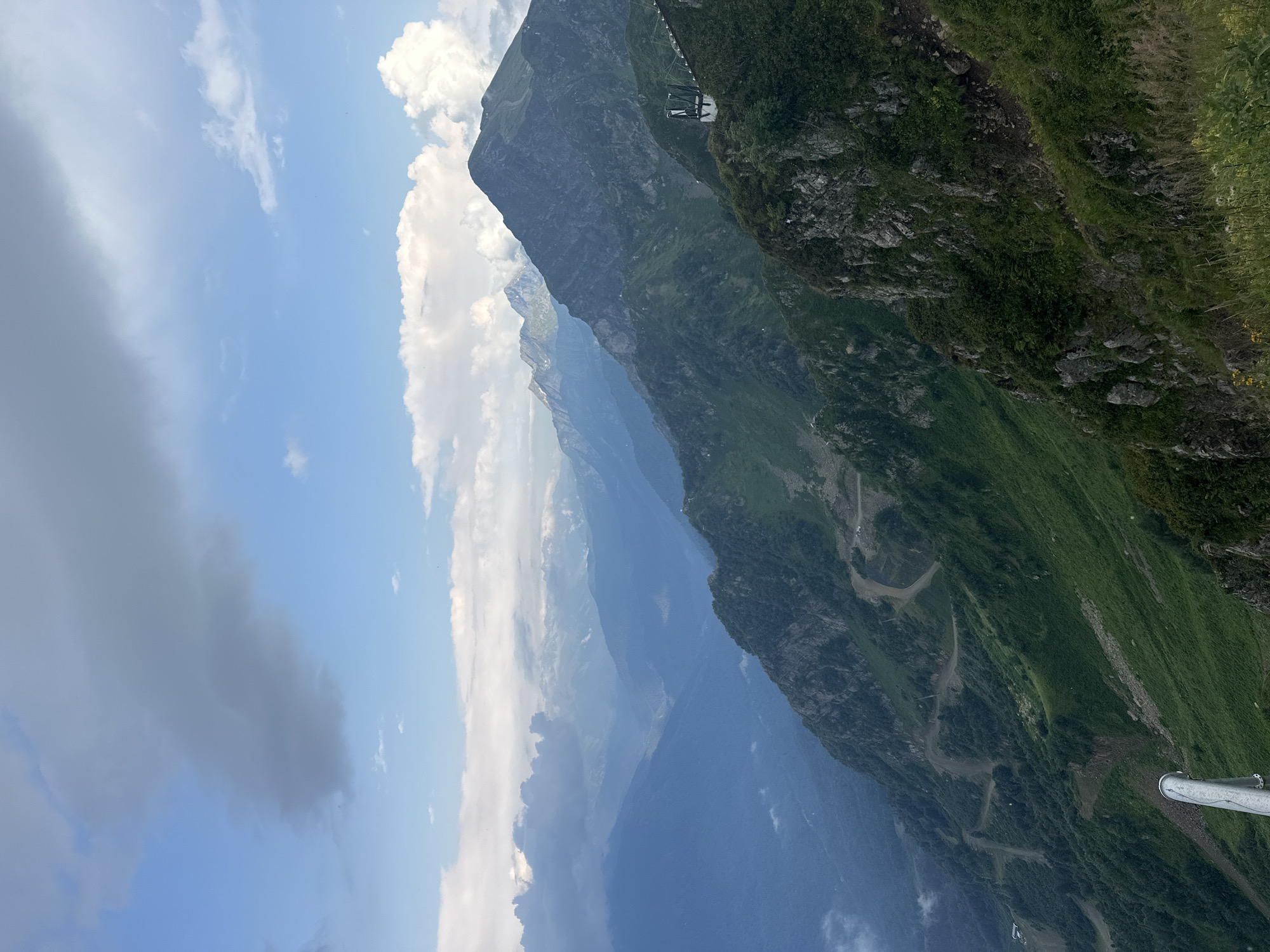 The image depicts a panoramic view of a mountainous landscape, likely taken from a high vantage point. The scene is characterized by rolling green hills and steep, rocky slopes, interspersed with patches of trees and vegetation. A winding road or trail is visible on one of the slopes, suggesting a popular hiking or scenic drive location. In the distance, layers of mountains recede into a hazy blue, hinting at a vast and deep valley. The sky above is a mix of clear blue and scattered clouds, with some larger, darker clouds suggesting approaching or passing weather. A white railing, possibly part of a viewing platform or pathway, is visible in the lower left corner, indicating human presence and infrastructure within the natural environment. A modern structure with a dark roof and white walls, resembling a ski lift station or observation tower, is perched on a ridge in the middle right of the image. This type of scenery is characteristic of mountain resorts, and given the context of Estosadok, Russia, it is plausible this image was captured in or around the ski resort areas there, such as Rosa Khutor or Gazprom Mountain Resort, known for their dramatic mountain vistas and extensive cable car systems.