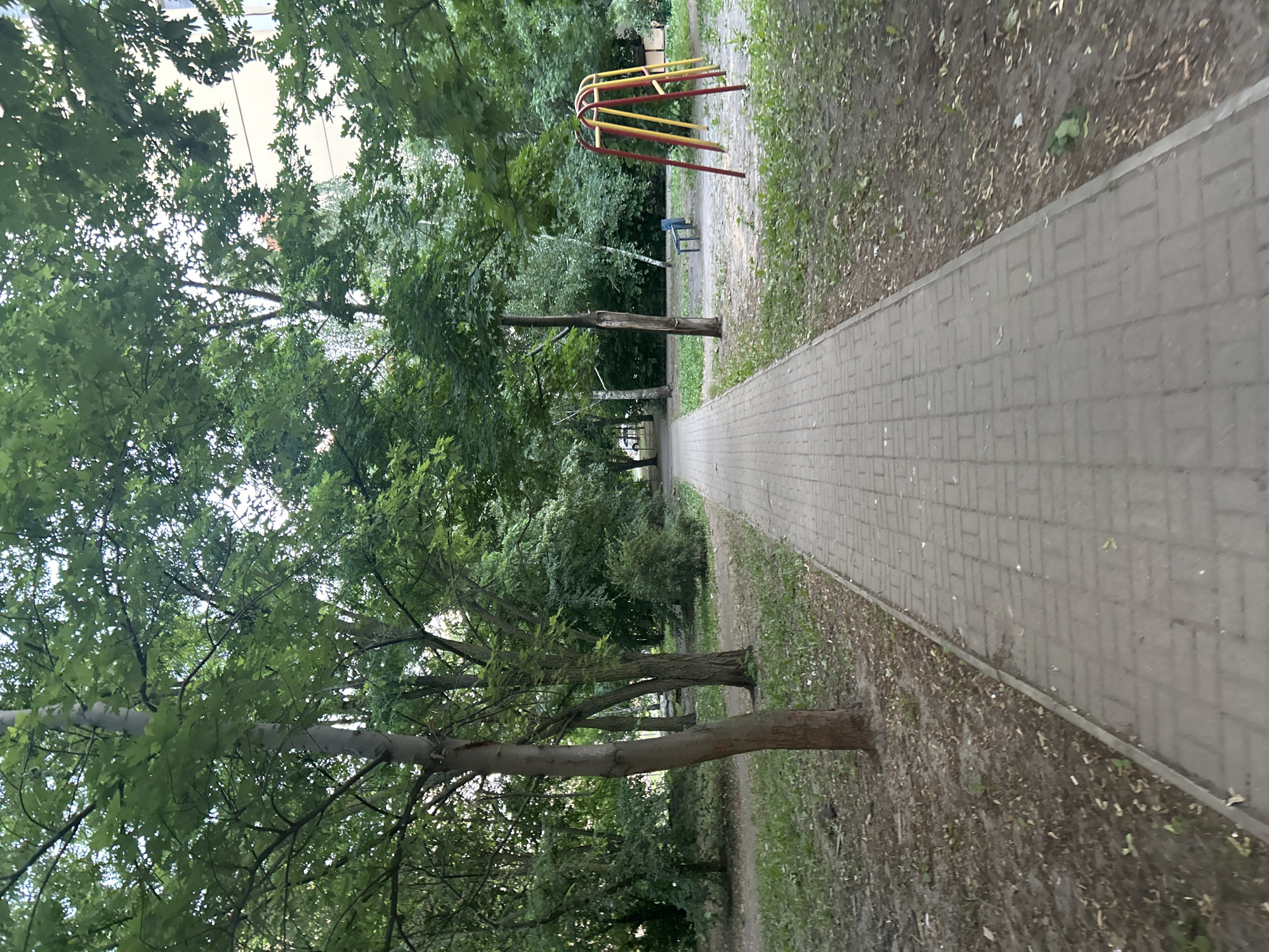 The image shows a paved walkway leading through a park-like area with mature trees and a playground. The walkway is made of grey rectangular pavers and is bordered by dry dirt and patchy green grass. The trees are lush with green leaves, indicating a warm season, likely summer. On the right side of the walkway, there is a children's playground with a yellow and red swing set and a blue bench. In the background, a building with a light-colored facade is visible through the trees. The overall scene depicts a typical urban park or courtyard in Novaya Usman, Russia. There are no people visible in the image.
