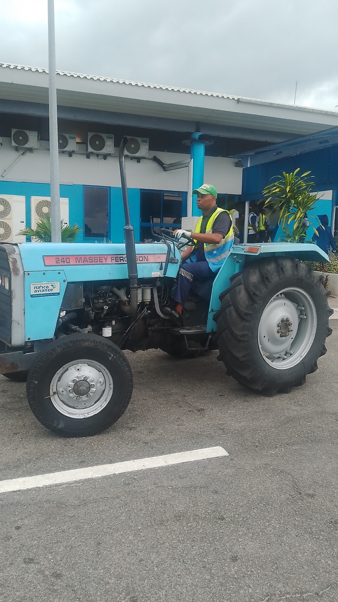 A man in a green cap and a yellow and blue reflective vest is operating a light blue Massey Ferguson 240 tractor on a paved surface in Orerokpe, Nigeria. The tractor has a metal exhaust pipe on the left side. The man is holding the steering wheel with gloved hands. Behind the tractor, a modern building with blue accents and multiple air conditioning units is visible. The sky is overcast. The tractor is positioned on a road marked with a white line.