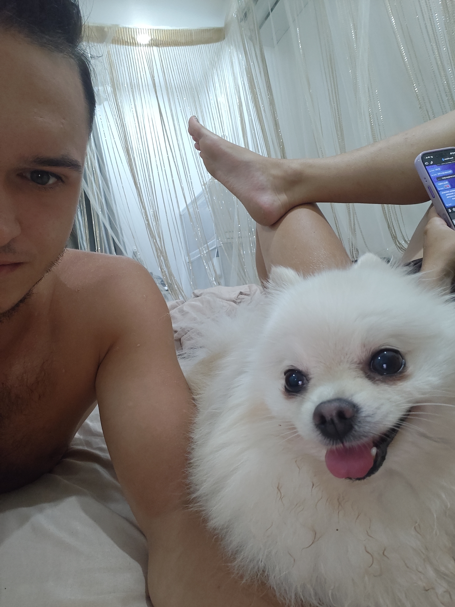 This media file captures a close-up, selfie-style shot featuring a person and a white Pomeranian dog. The person, partially visible, is lying down, with their face in the foreground on the left, looking towards the camera. They appear to have dark hair pulled back, and their shoulder and upper arm are also visible. The dog, positioned in the center right, is also lying down and looking directly at the camera with its tongue slightly out, conveying a happy or excited expression. The dog's fur is fluffy and white.
The background consists of draped sheer fabric with vertical strings or embellishments, creating a soft, curtain-like effect. The lighting suggests it is daytime, possibly indoors, with light filtering through the fabric. A foot is visible in the upper right quadrant, suggesting the person's legs are extended. A mobile phone, held by the person's other hand (partially visible in the upper right), displays a screen with text and icons, though the content is not clearly discernible. The setting appears to be a relaxed, indoor environment, possibly a bed or sofa, indicated by the soft fabric. The overall mood is casual and affectionate, highlighting the bond between the person and their pet. No specific time of day or weather conditions are discernible beyond the implied daylight. The location context provided places the scene in Tay Nha Trang Ward, Vietnam.