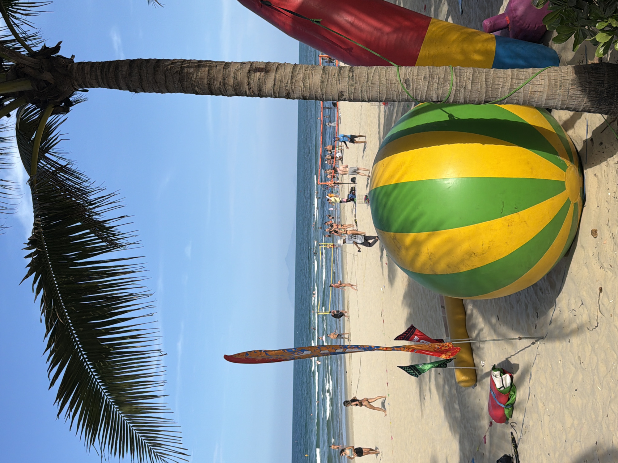 The image depicts a beach scene in Đà Nẵng, Vietnam. The beach is sandy and populated with numerous people sunbathing, swimming, and walking along the shoreline. In the foreground, there is a section of a palm tree trunk with leaves and various inflated beach toys including a large yellow and green striped ball. A long kite with small flags is on the sand. The sea is visible in the middle ground, with gentle waves reaching the shore. The sky is a clear blue, and sunlight appears to be casting shadows.