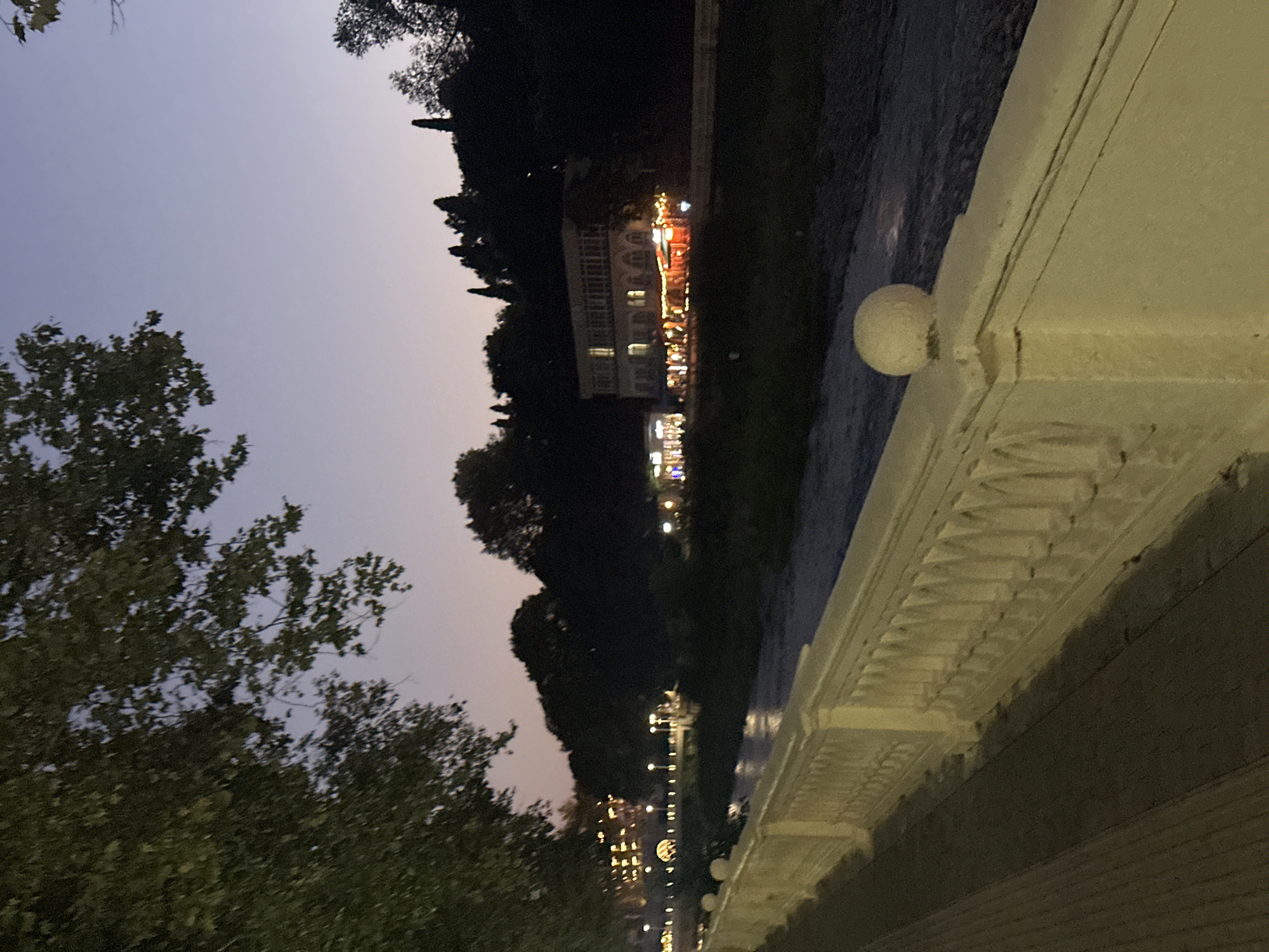 The image depicts a riverside embankment at dusk in Sochi, Russia, likely in the vicinity of the central district. On the left, a tree with dense foliage casts shadows. A paved walkway runs alongside a white, ornate balustrade. Beyond the balustrade, a river flows, its surface reflecting the dim light. Across the river, a dark hillside is covered with trees.
On the hillside, several buildings are visible, illuminated by warm, yellow lights. One prominent building, a multi-story structure with arched windows and a classical facade, appears to be a restaurant or hotel, with outdoor seating illuminated by string lights. Further to the left, another building with similar lighting is partially visible. Streetlights line the embankment across the river, casting pools of light. The sky above is a gradient of deep blue and purple, characteristic of twilight. No people are clearly discernible in the image. The scene suggests a tranquil evening ambiance.