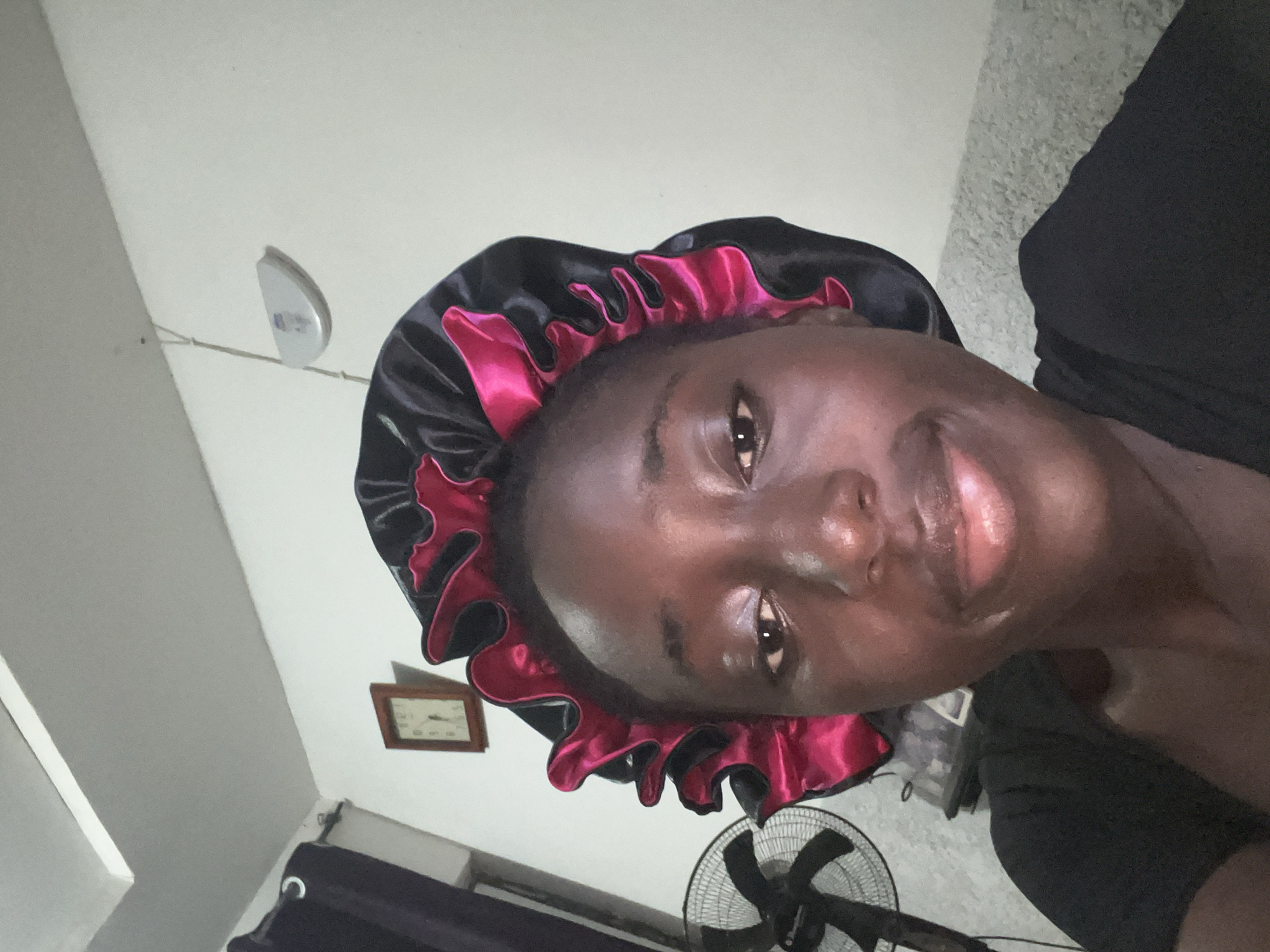 A person is visible, wearing a black and pink bonnet. The location seems to be indoors, possibly a bedroom, as indicated by the presence of a fan, a wall clock, and electrical fittings on the wall. The picture does not confirm the location in Abuja, Nigeria.