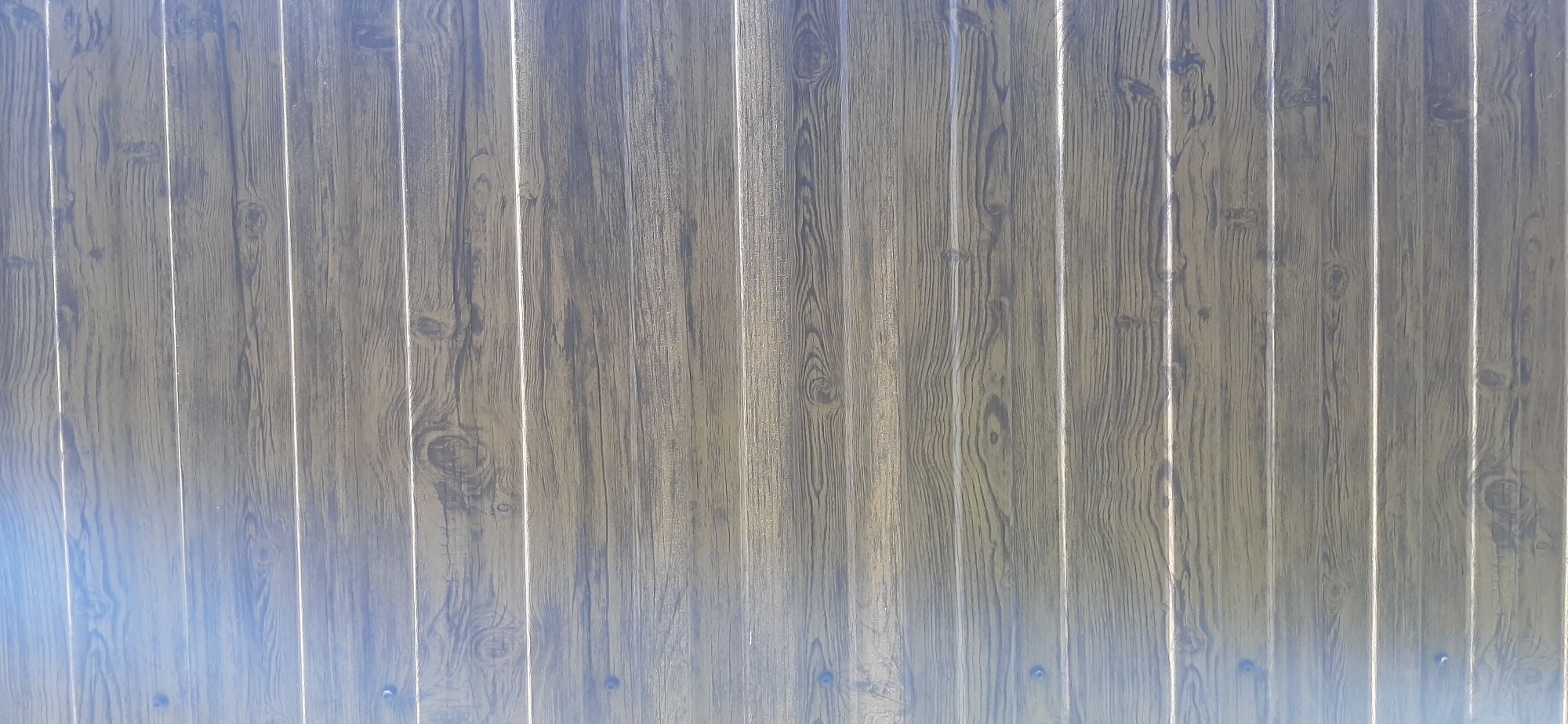 The image shows a close-up, full-frame view of a corrugated metal fence with a faux wood grain pattern. The vertical planks are a muted brown and grey, with lighter grey lines simulating wood grain and knot details. The surface appears to be outdoors, as evidenced by a subtle blueish light cast on the lower left portion of the fence, suggesting sky reflection. There are no people, animals, or other objects visible in the frame. The setting is a generic outdoor environment, with no specific landmarks or details that would indicate its location within Yessentukskaya, Russia, or any other city.