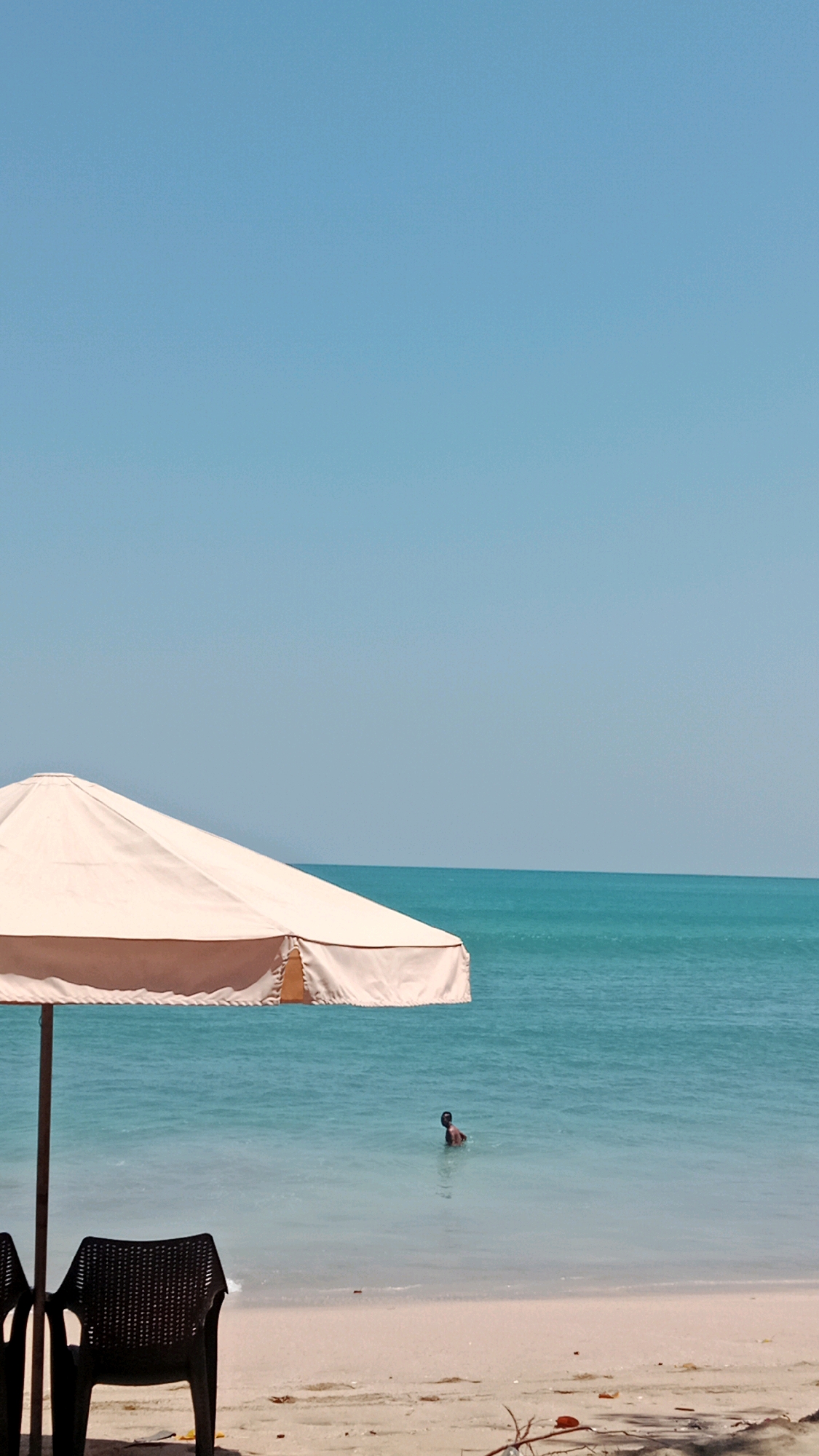 The image depicts a serene beach scene at Kuta, Indonesia. A light-colored beach umbrella is prominently positioned in the foreground on the left, casting a shadow. Two dark plastic chairs are placed in front of the umbrella. The gentle waves of the turquoise ocean are visible in the midground, with a solitary person wading in the water, their upper body and head above the surface. The sandy beach stretches across the bottom of the frame. The sky above is a clear, bright blue, indicating a sunny day.