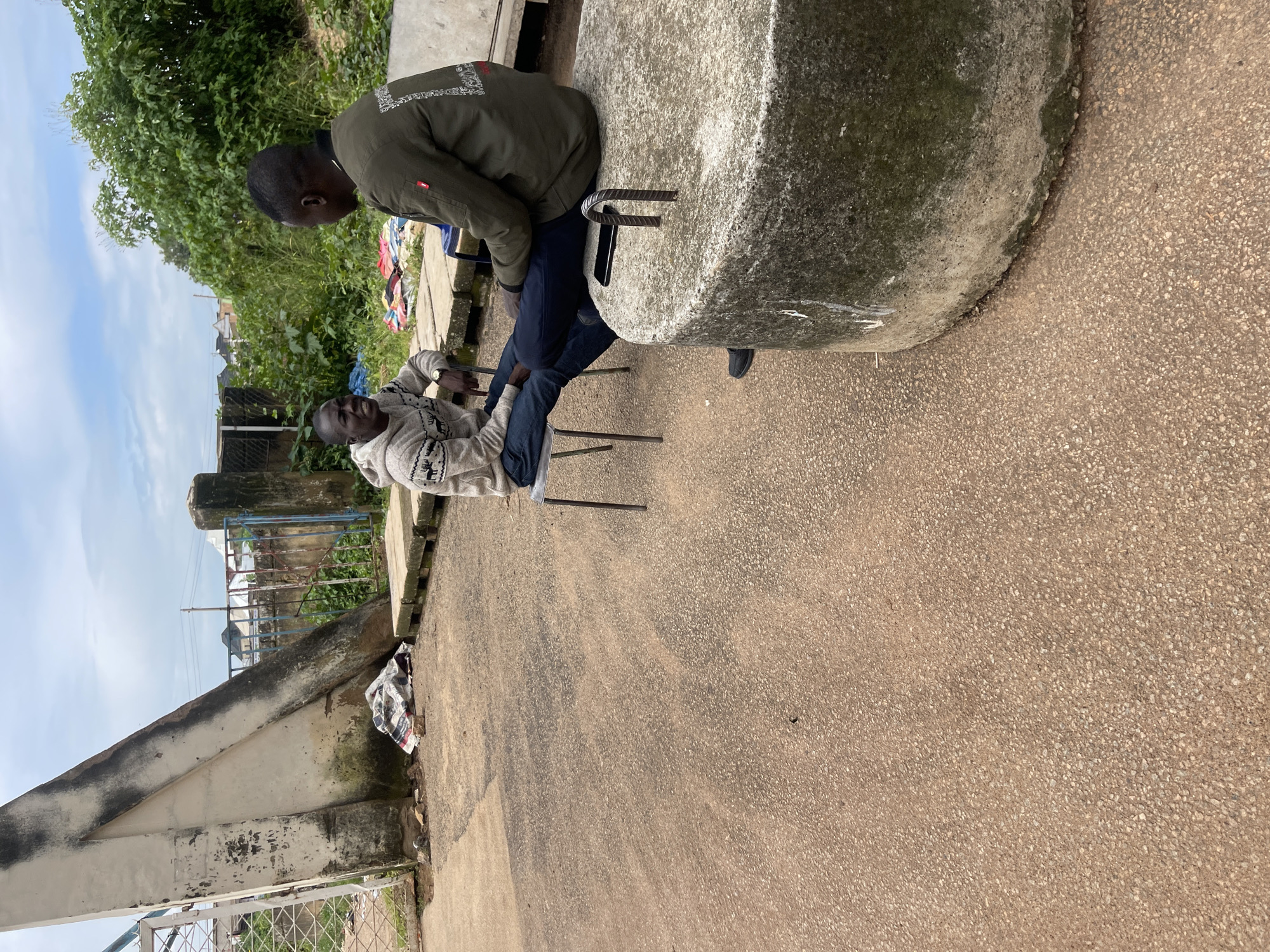 Two young Black men are sitting outdoors in an urban environment, with one man on a metal chair and the other on a large, round concrete object. The man on the chair, wearing a grey and white patterned hoodie and blue jeans, is looking towards the other man with an open mouth, appearing to be speaking or laughing. He has a watch on his left wrist. The man on the concrete object is facing away from the camera, wearing a dark green jacket with white lettering on the back and dark pants. His arm is resting on the concrete object. Between the two men, on the concrete object, is a black smartphone. The ground is paved with asphalt. In the background, there is a large, white, triangular concrete structure to the left, a metal fence, some overgrown green vegetation, and buildings in the distance. The sky is overcast with clouds. The image appears to have been taken in Rafin Par, Nigeria, based on the provided context.