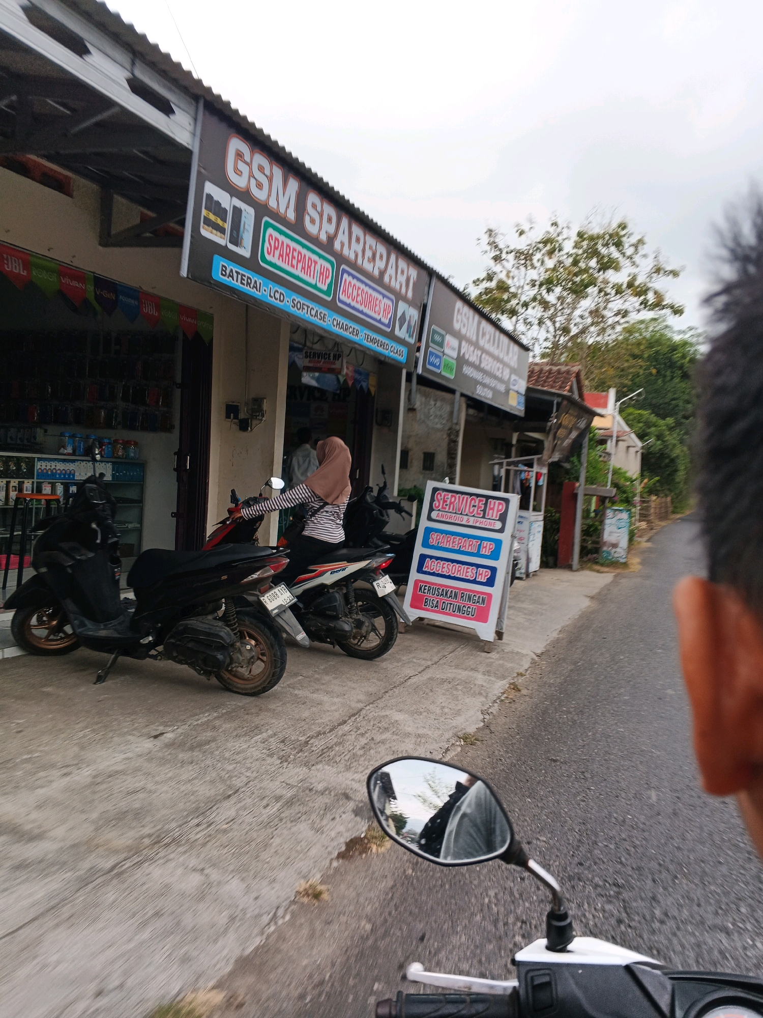 A street-level, outdoor shot captures a mobile phone repair and accessories shop. In the foreground, two motorcycles are parked on the sidewalk. A woman wearing a hijab and a striped shirt is seated on one of the motorcycles, facing the shop. A sign for "GSM SPAREPART HP" is prominently displayed, listing services like "BATTERAI - LCD - SOFTCASE - CHARGER - TEMPERED GLASS". Another sign advertises "SERVICE HP" for Android and iPhone, with a note that minor repairs can be done while waiting. The shop appears to be a small, local business situated along a road with trees and other buildings in the background. The overall scene suggests a typical day at a neighborhood mobile phone service center.