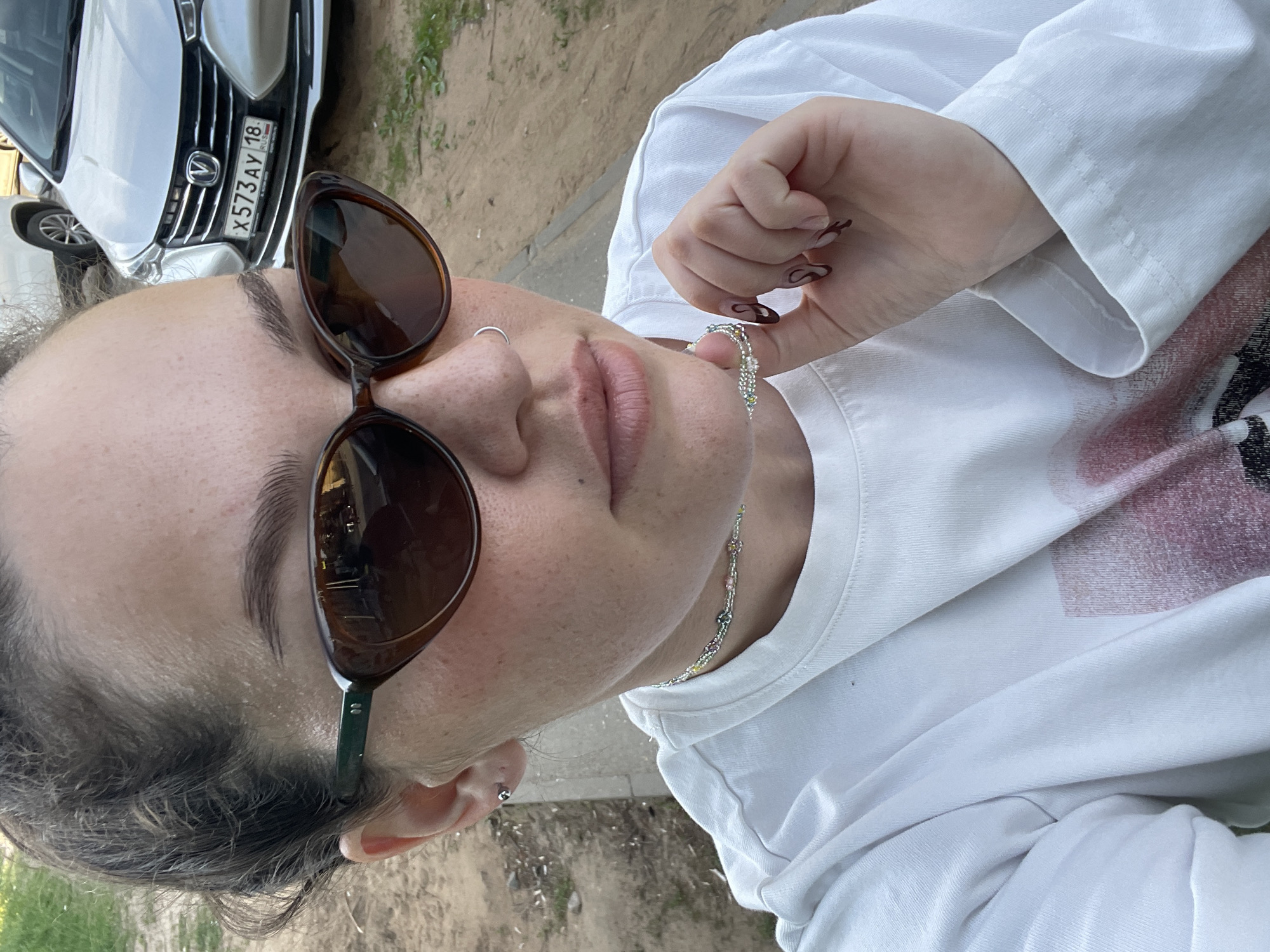 A woman in sunglasses and a white shirt is depicted in a close-up, rotated image. Her right hand holds a beaded necklace. The license plate X573AY18 is visible on a car, indicating Izhevsk, Russia. The environment suggests an outdoor, possibly urban setting.