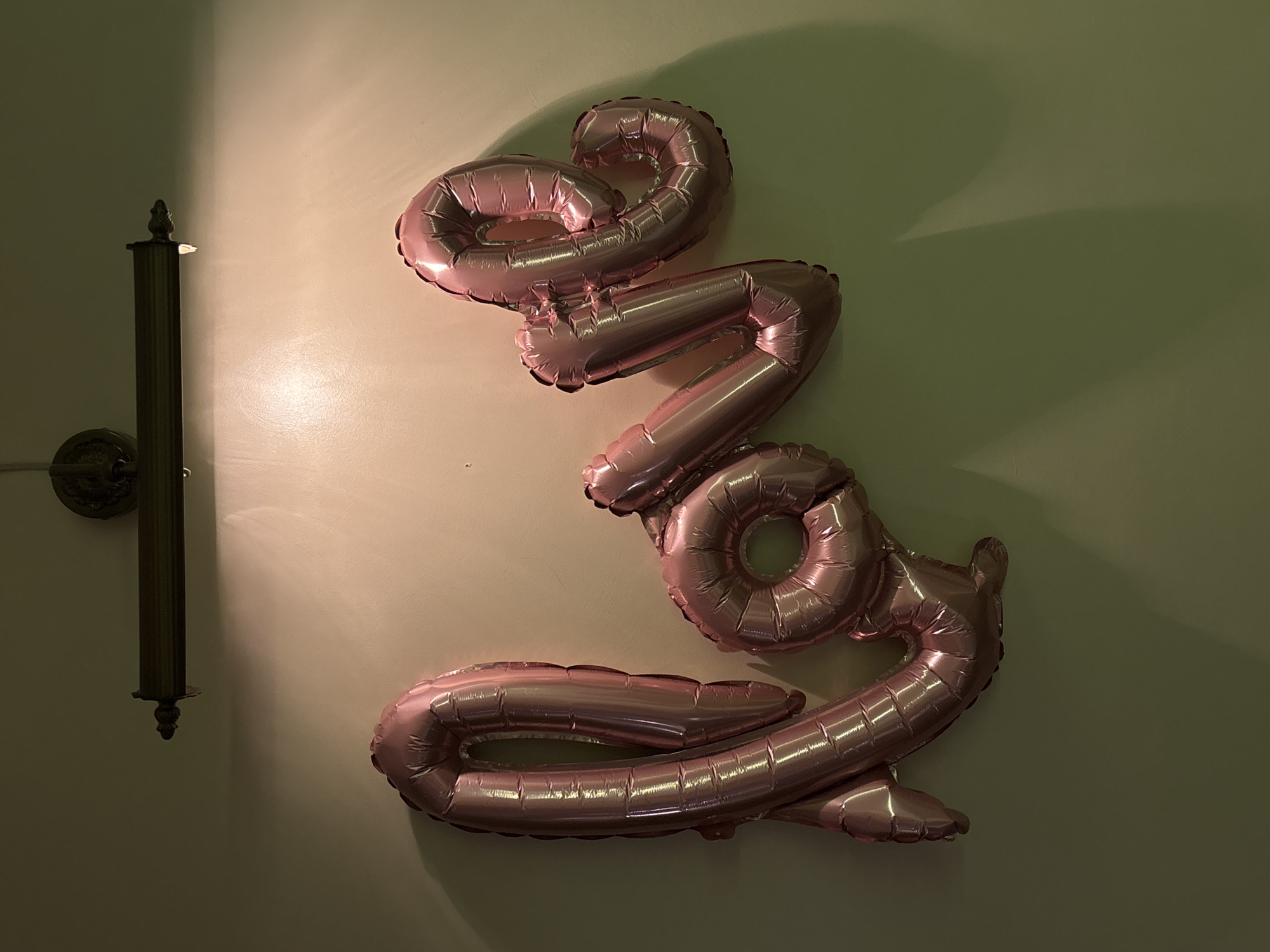 The image shows a wall with rose-gold foil balloons forming the number "2023." A sconce-style light fixture is mounted on the wall to the left of the balloons. The wall is light-colored and unevenly lit. The scene appears to be indoors. The location in Krasnoyarsk, Russia is not discernible from the image.