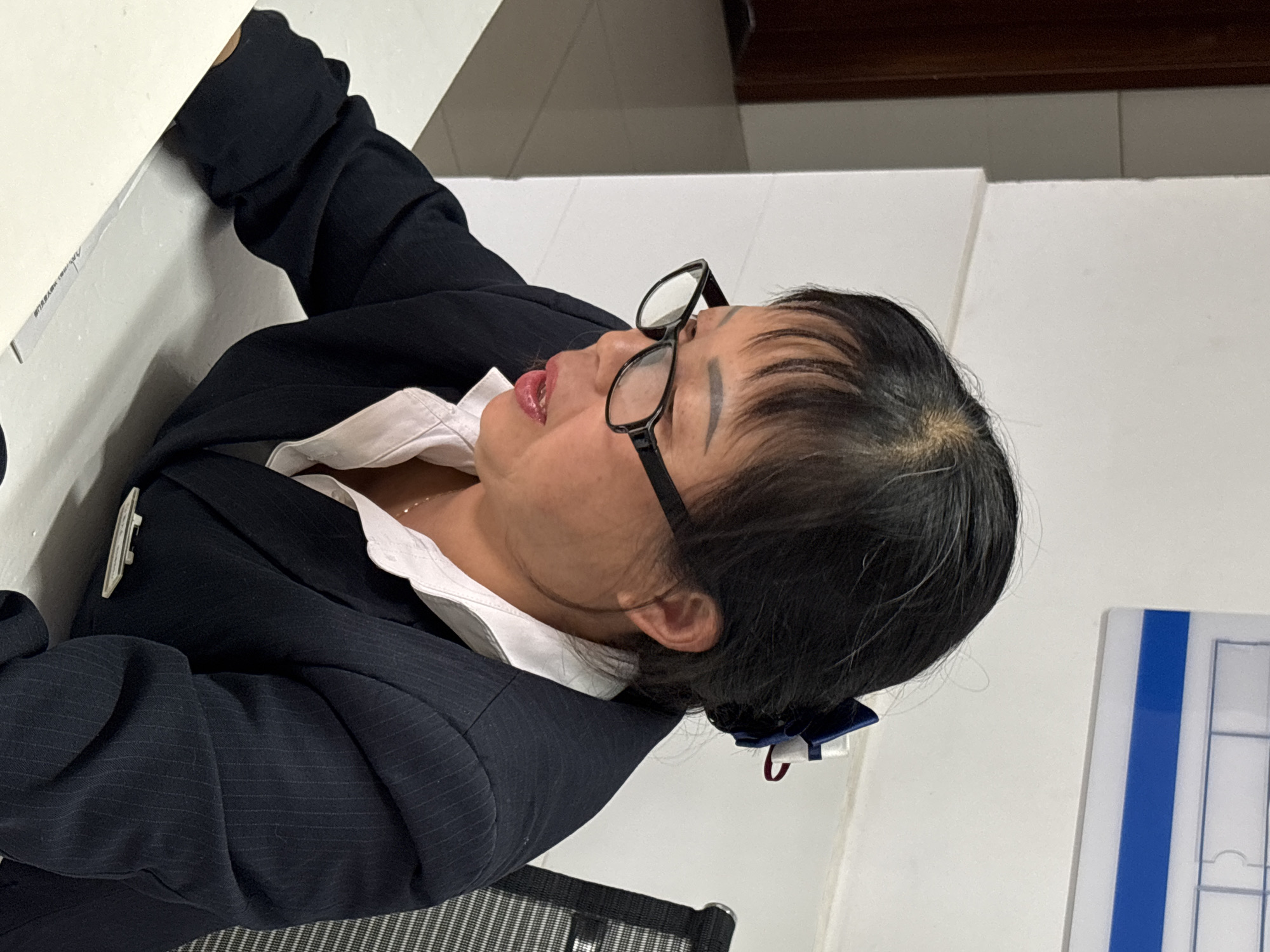 A woman wearing glasses, a white collared shirt, and a navy blue pinstripe suit jacket is visible. The woman is leaning back, seemingly at a desk. A white and blue object is visible on the right, and what appears to be a desk and office chair are visible on the bottom of the frame. The location is Jiangjin, China.