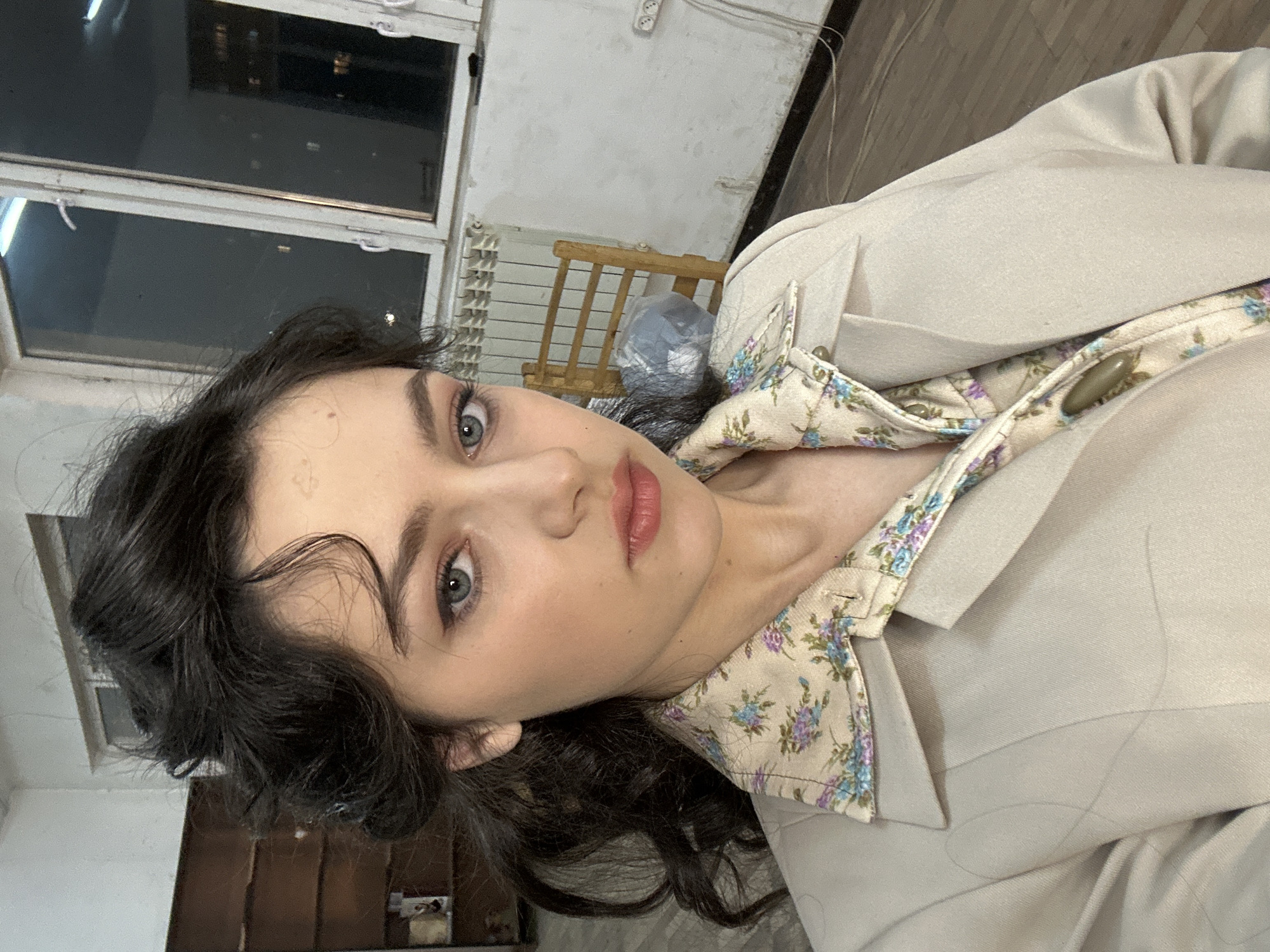 A woman with dark, wavy hair and blue eyes is the focal point of the image. She is wearing a light-colored blazer over a floral-patterned shirt. The blazer is partially buttoned. She has fair skin and is wearing lipstick.
The background includes a window with multiple panes, revealing an urban landscape, indicating a city location. There is a radiator and a wooden chair behind her. Other furniture and items are visible in the background, suggesting an indoor setting, possibly an apartment or studio.
Based on the stated location, the city is Yerevan, Armenia, but specific details that would uniquely identify the scene as being in that location are not present. The image is primarily a portrait of the woman.