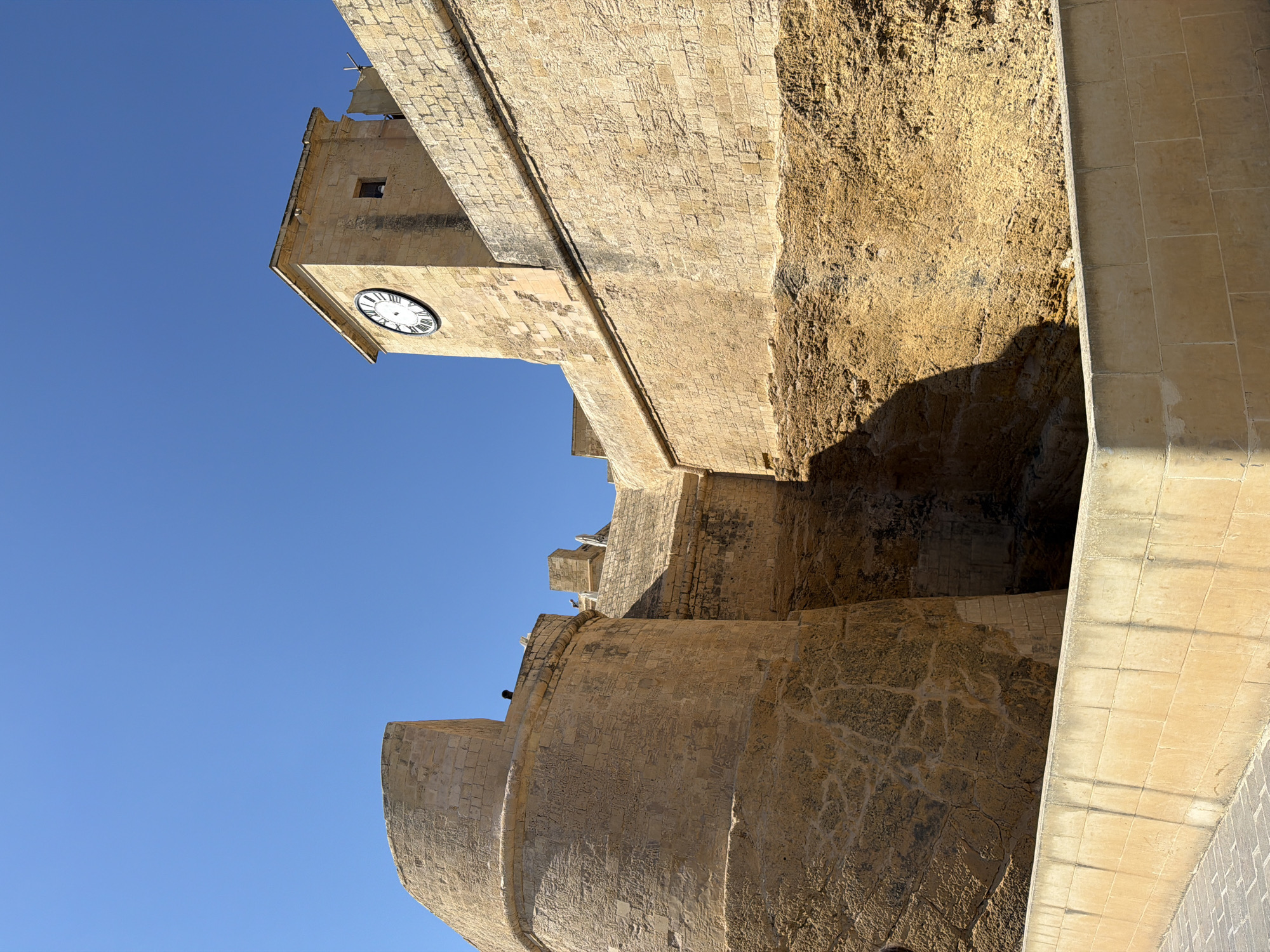 The image captures a portion of the Cittadella in Victoria, Gozo, Malta. The architecture is predominantly stone, with warm, sandy-colored walls built from large, rectangular blocks. In the foreground, a lower wall, also constructed of stone blocks, lines a pathway paved with patterned bricks. This wall exhibits some discoloration and shadowing, suggesting it's a well-trodden area.
Dominating the mid-ground and background are the imposing fortifications of the Cittadella. A large, curved bastion with a weathered texture and visible stone seams occupies the left side of the frame. To its right, a taller, square clock tower stands out against a clear, vibrant blue sky. The clock face is circular with Roman numerals and visible hands, indicating the time. Adjacent to the clock tower, a long, high wall extends horizontally, also made of stone blocks and showing signs of age. The base of this wall appears to be built into the natural rock face, which is rough and textured, with crevices and shadows.
Two figures are visible on top of the left bastion. One is partially obscured and standing near the edge, while the other is a silhouette further along the parapet. The scene is bathed in bright sunlight, casting sharp shadows that emphasize the textures and forms of the ancient stonework. The overall impression is one of historical grandeur and architectural solidity under a clear, sunny sky.