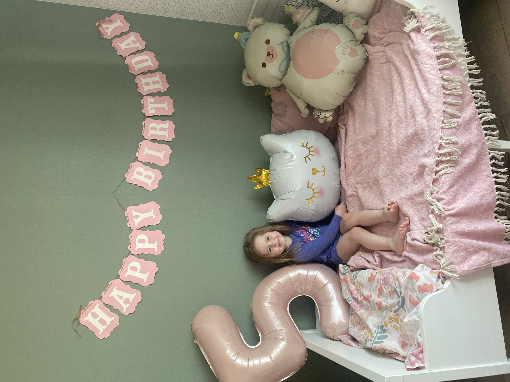A girl lies on a bed adorned with pink blankets and balloons, including a "5" balloon, beneath a "Happy Birthday" banner on a green wall. The scene is indoors, likely a child's bedroom. The location, Izhevsk, Russia, is provided as context but not visible in the image.