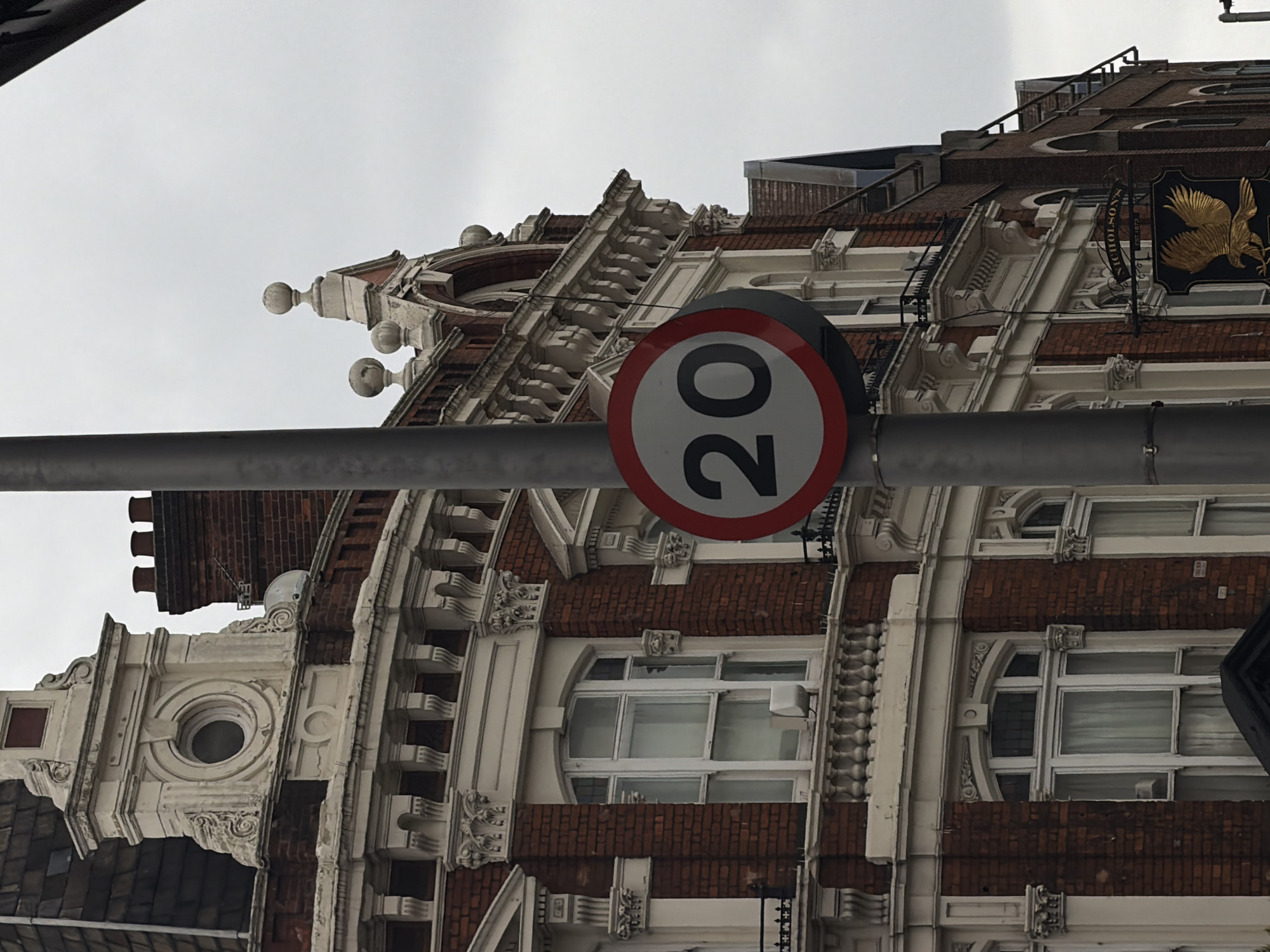 The image shows a building exterior, likely in London, United Kingdom. A "20" speed limit sign is prominently featured, attached to a metal pole. Behind the sign is a multi-story building with brick and white architectural details. The upper part of the building has elaborate decorations. A sign is visible on the right side of the building featuring a golden eagle and the name "NICHOLSON'S". The sky is overcast.