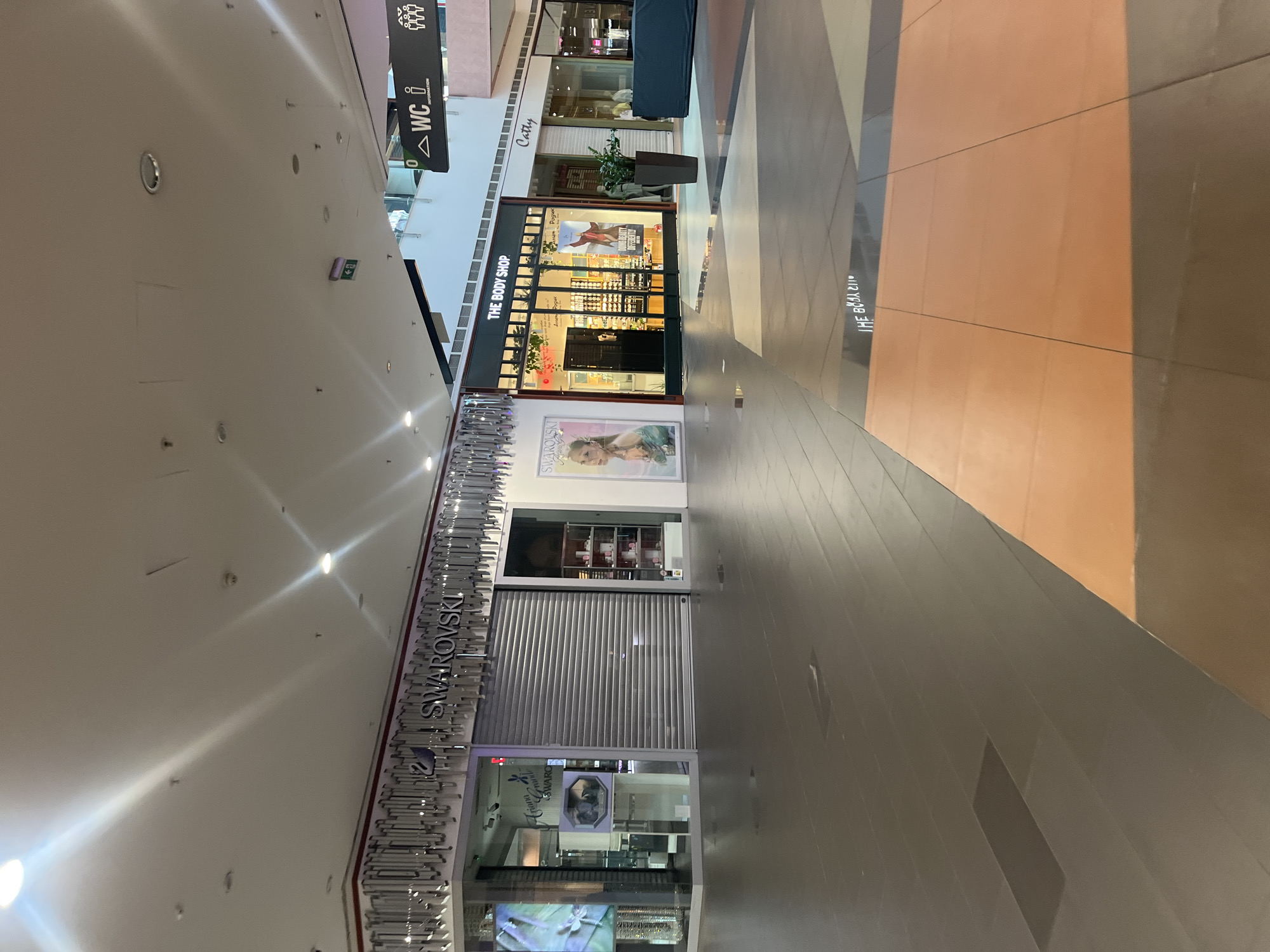 The image depicts the interior of a shopping mall, likely located in Sofia, Bulgaria. Focus is on a corridor displaying closed stores with their metal shutter doors pulled down. Among them are a Swarovski shop on the left, recognizable by the brand's name displayed above, and The Body Shop further down the corridor, clearly identifiable by its signage. Other retail establishments are also visible. The floor features a mix of gray and orange tiling, and the ceiling shows rows of recessed lights. An overhead sign with the "WC" (toilet) symbol is seen, indicating restroom facilities. The general impression is that the mall is either closed or experiencing limited operation. No people are visible in the scene.