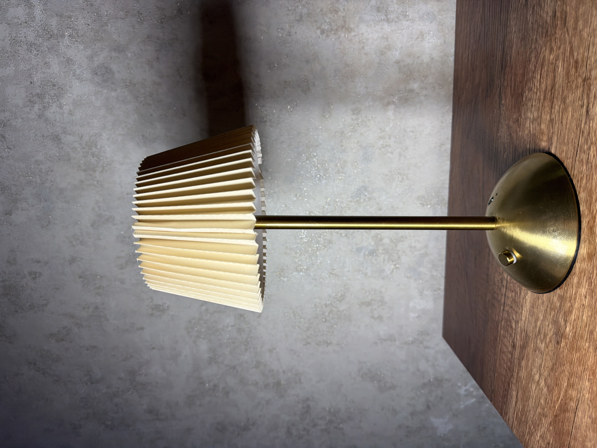 A brass table lamp with a pleated cream-colored lampshade stands on a dark brown wooden surface. The lamp has a rounded base with a small brass knob or switch on its side, and a slender brass stem supporting the shade. The background is a textured gray wall, which appears to be concrete or stucco, with subtle variations in color and tone. The lighting casts a shadow of the lampshade onto the wall behind it. The image does not contain any people or animals, and no specific city or location is depicted or identifiable.