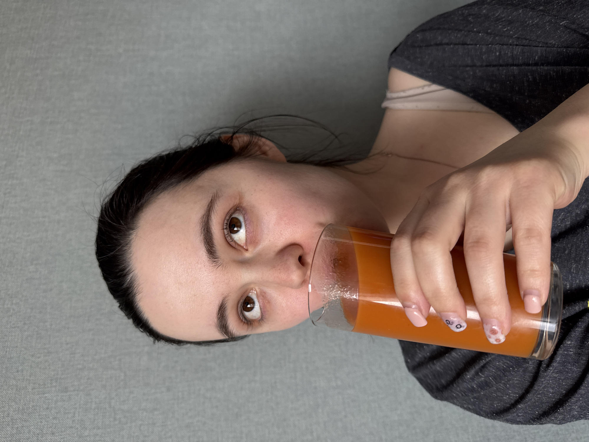 A woman is drinking from a glass of orange juice. She is wearing a gray shirt and has dark hair pulled back. Her fingernails are painted with a light pink color and small floral designs. The background is a plain gray wall. There is no evidence to suggest that the location is Izhevsk, Russia.