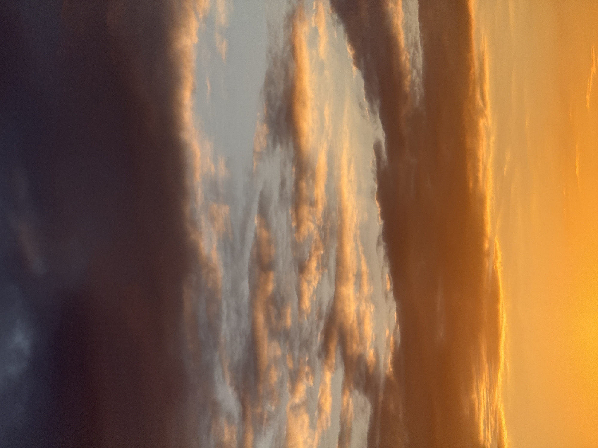 The image, oriented vertically, captures a dramatic sky filled with clouds, indicative of either sunrise or sunset. The right side of the frame is bathed in brilliant golden and orange light, suggesting the low position of the sun just beyond the view. This warm illumination intensely highlights the edges and undersides of substantial cloud formations, creating vibrant glows and distinct outlines against a softer, lighter blue-grey sky behind them.
As the eye moves leftward, the clouds gradually transition into deeper, more subdued tones of grey and purple-blue, indicating areas of shadow and less direct sunlight. The far left portion of the image is dominated by a dark, almost charcoal-colored cloud mass, providing a strong contrast to the luminous right side. The clouds exhibit varied textures and depths, from soft wisps to more solid, volumetric structures catching the light.
There are no visible people, man-made objects, or text. The scene is a powerful display of natural atmospheric light and shadow, creating a sense of awe. The weather appears to be partly cloudy. While the location context of Perm, Russia is provided, the image itself does not contain specific geographical cues.