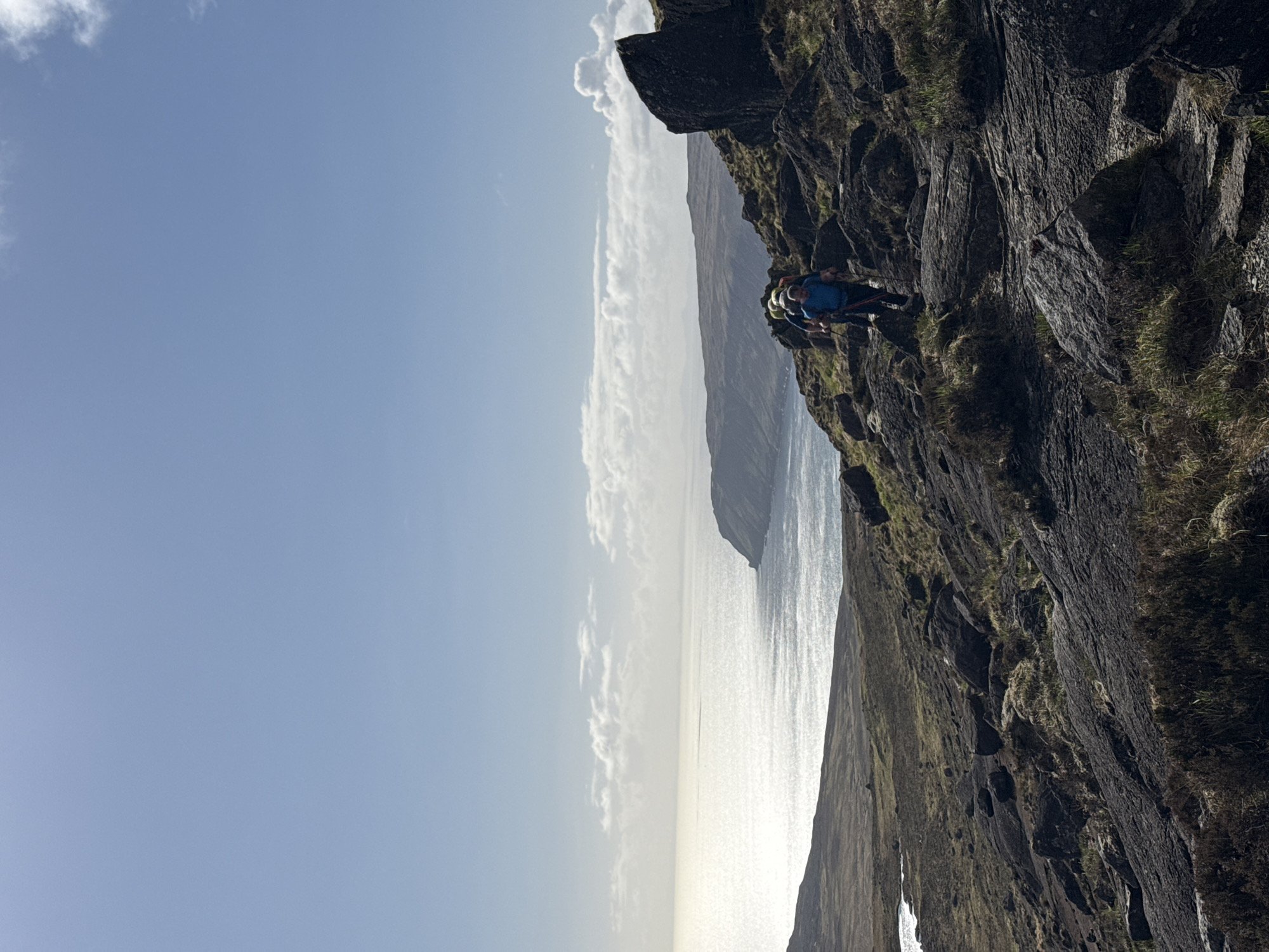 A climber is scaling a rock face on a cliff overlooking the ocean. The sky is mostly clear with some clouds. The rock face is dark and textured with some patches of green vegetation.