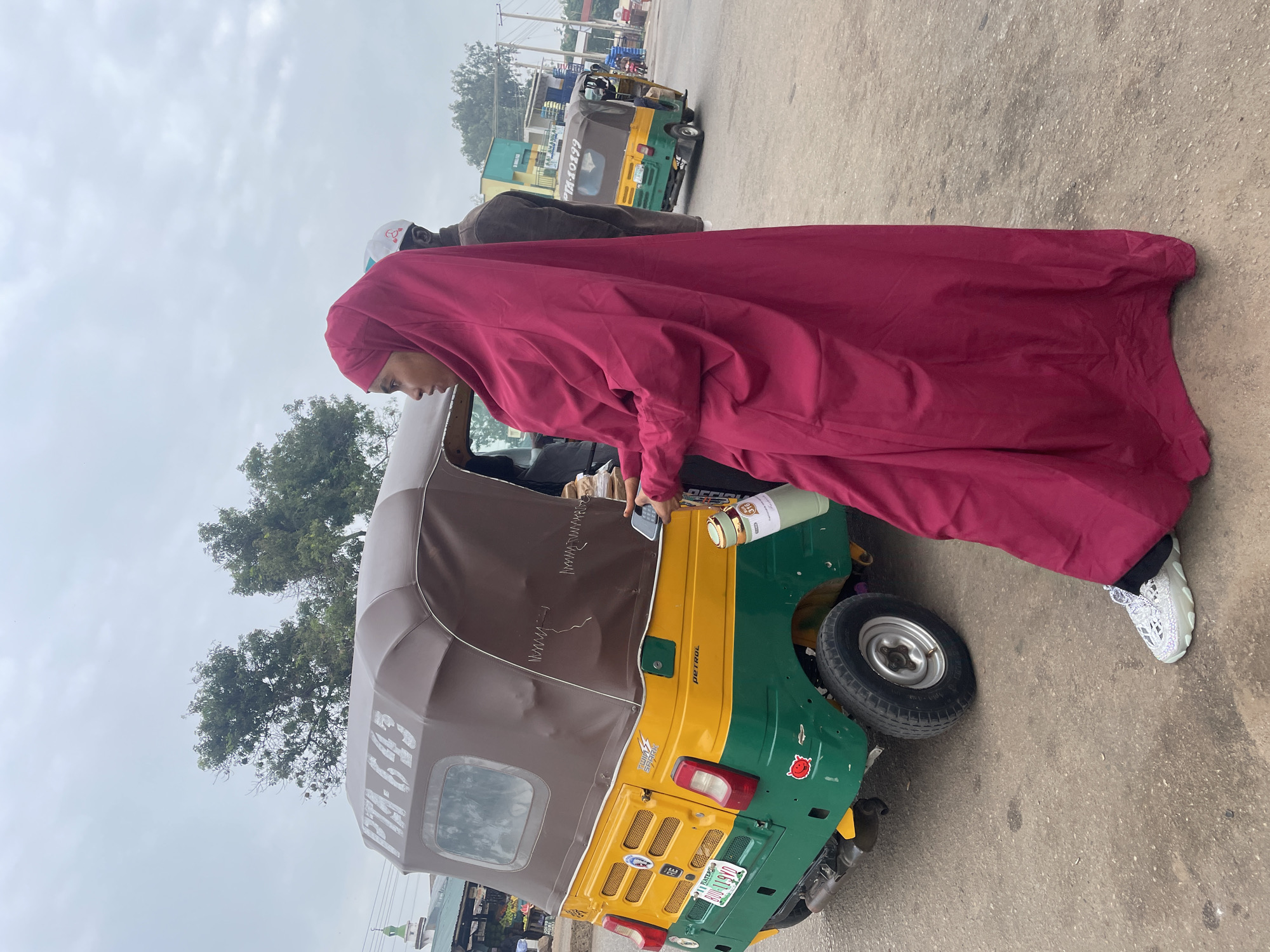 A full-shot, eye-level, outdoor photograph depicts a woman in a bright pink abaya and hijab standing next to a yellow and green auto-rickshaw with the license plate "BOG 119 VIO". The auto-rickshaw has a brown canopy with the inscription "PTA - 643" visible on the side. The woman is looking to her left, holding a smartphone in her right hand and a reusable water bottle in her left. A second person, wearing a brown jacket and a white cap, is partially visible behind her, also looking to the left. The auto-rickshaw is parked on a paved road, possibly in a market or transportation hub, with other vehicles and buildings visible in the background, suggesting an urban environment in Jos, Nigeria. The sky is overcast.