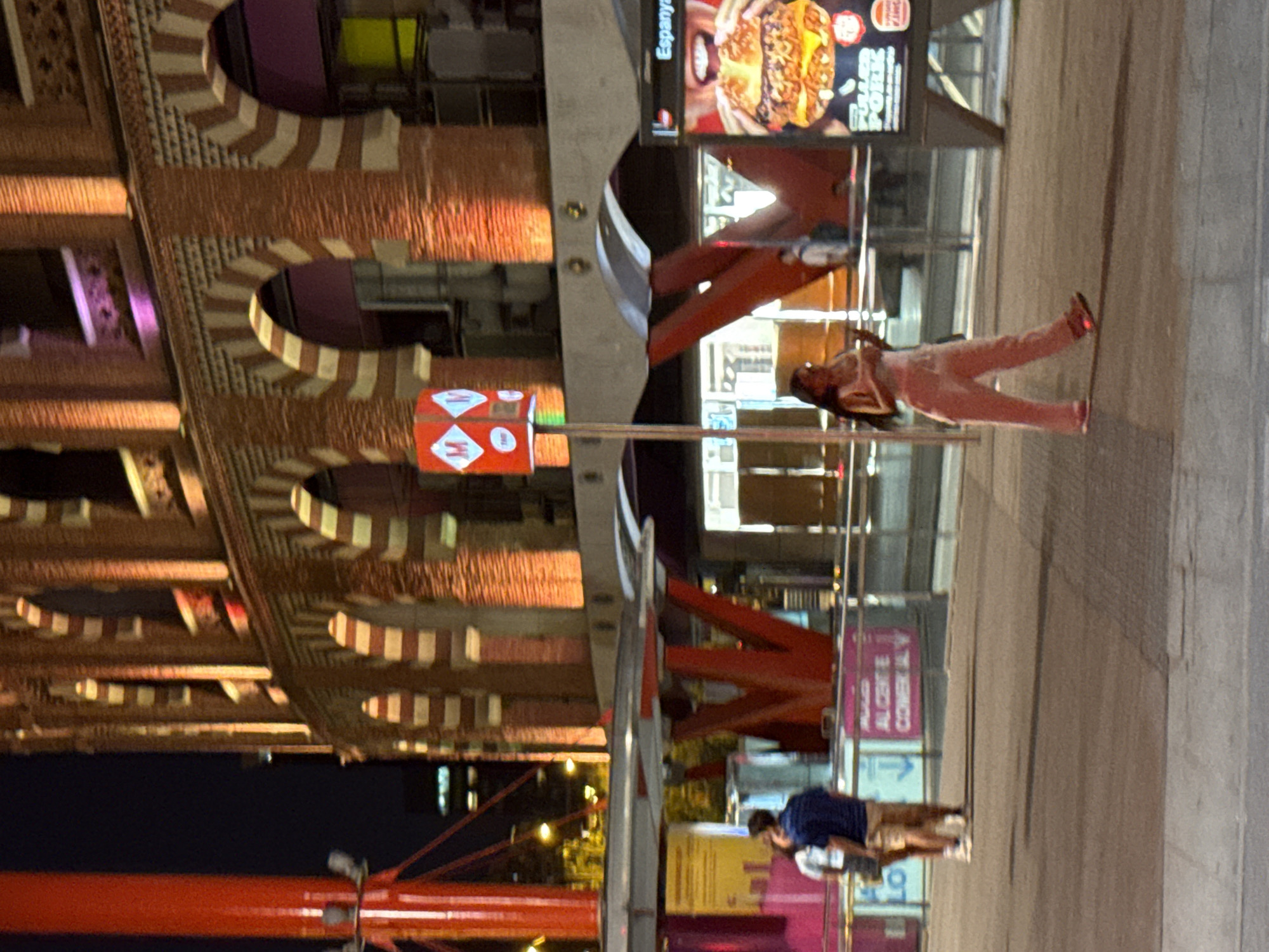 In Barcelona, Spain, the image captures a nighttime scene at the Las Arenas de Barcelona, a former bullfighting arena converted into a shopping center. In the foreground, a woman in a light-colored outfit walks across a paved area, holding a phone. Behind her, a couple stands near a railing, facing each other. The iconic circular building of Las Arenas is prominently featured in the background, illuminated with warm orange and purple lights, showcasing its distinctive arched windows. A red, pillar-like structure with metallic accents stands to the left of the frame, and a brightly lit advertisement for a burger is visible on the right. The overall atmosphere suggests a lively urban environment at night.