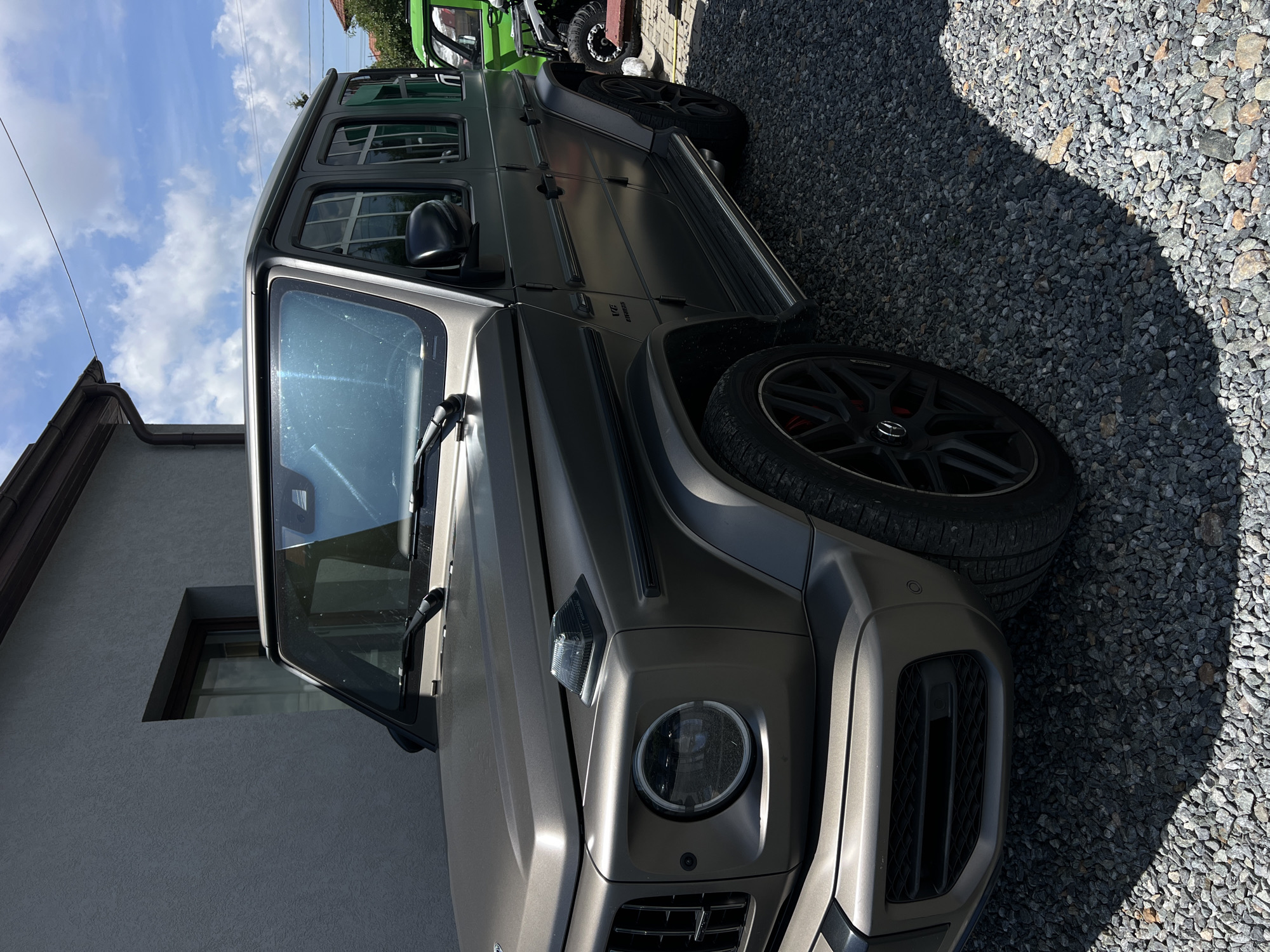 The image shows a matte gray Mercedes-Benz G-Class parked on a gravel surface outside a building. The building has a gray, textured exterior wall and a dark brown gutter. The sky is blue with white clouds. To the right of the G-Class, a green vehicle and a black ATV are partially visible. The scene appears to be in a residential or commercial area. The exact location within Cluj-Napoca, Romania cannot be determined from the visual information provided.