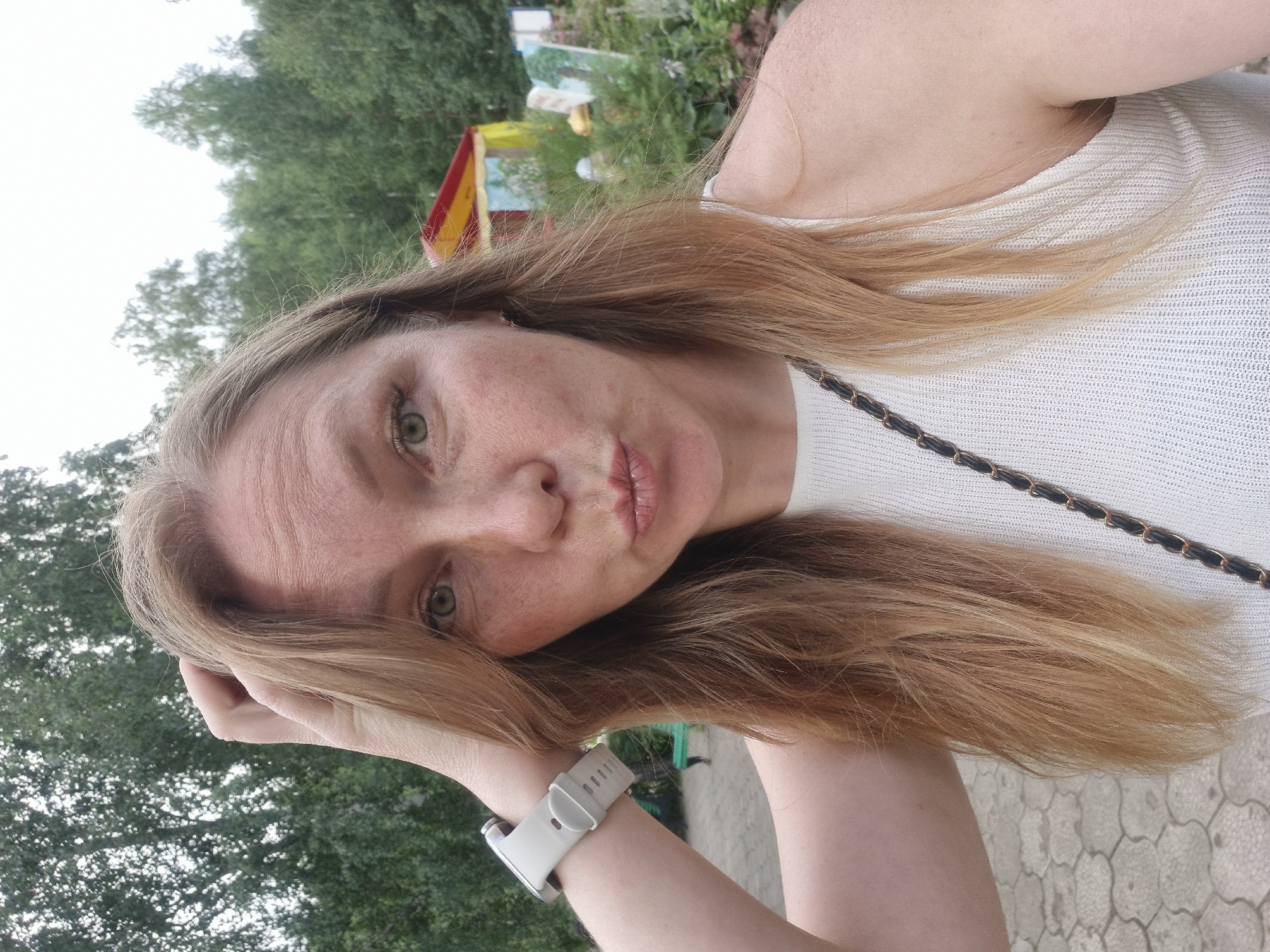 A woman with light brown hair and fair skin, wearing a white sleeveless top and a light grey smartwatch, is taking a selfie outdoors. She is puckering her lips and has her right hand raised to her head. In the background, there are trees, a paved walkway, and a colorful structure with a red and yellow roof, possibly a tent or a small building. The setting appears to be a park or outdoor recreational area in Chaikovsky, Russia.
