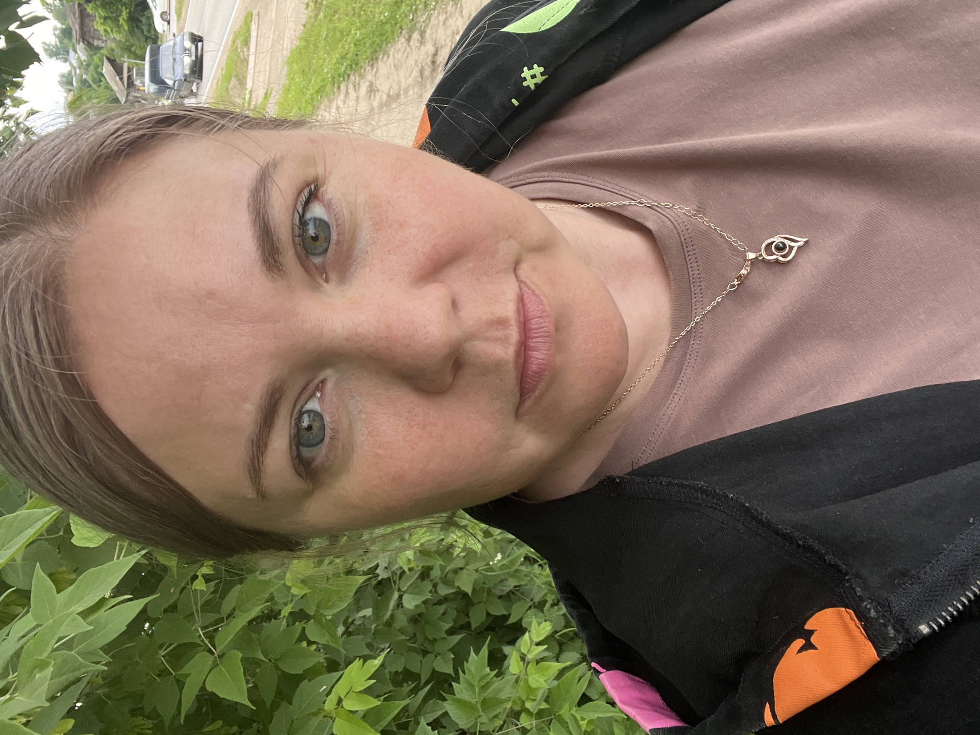 A woman is taking a selfie outdoors, with green foliage and a road visible in the background. She has light brown hair, blue-green eyes, and is wearing a light brown t-shirt and a black jacket with colorful accents. A silver necklace with a heart-shaped pendant is visible. The scene appears to be in a rural or suburban area, possibly near the city of Fokino, Russia, given the context. The road suggests a transportation route, and the presence of trees and vegetation indicates an outdoor setting.