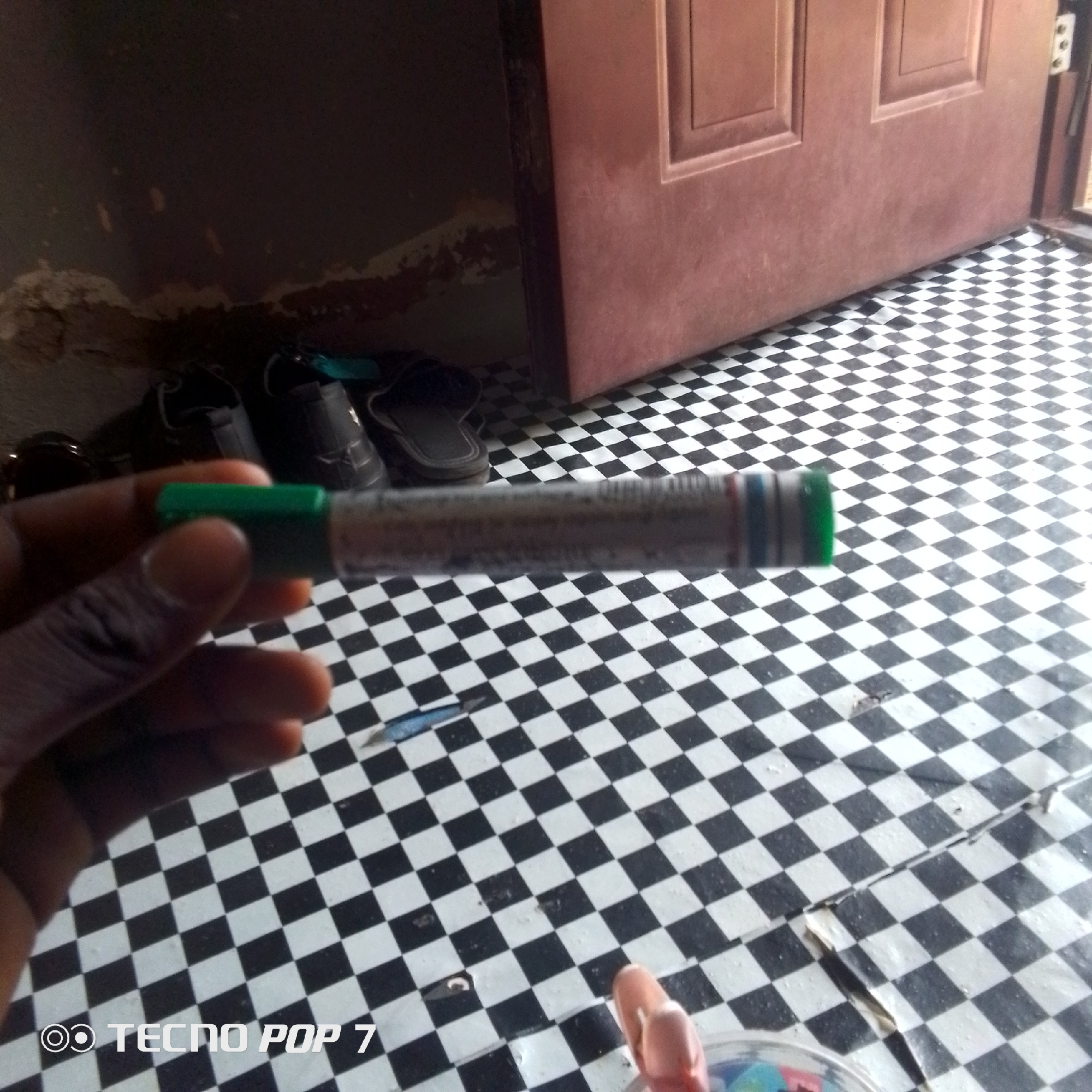 The image shows a close-up of a person's hand holding a pen-like object with a green cap. The pen has a metallic body with some markings and colored bands. The background is a black and white checkered floor, which appears to be indoors. To the left of the pen, a few pairs of shoes and sandals are visible, suggesting a domestic setting or an entrance area. In the background, a portion of a red-brown door is visible, with some textured details. The lighting suggests it could be daytime, possibly indoors with some natural light. The overall scene is casual and everyday. The bottom left corner of the image contains the text "TECNO POP 7" with a logo.