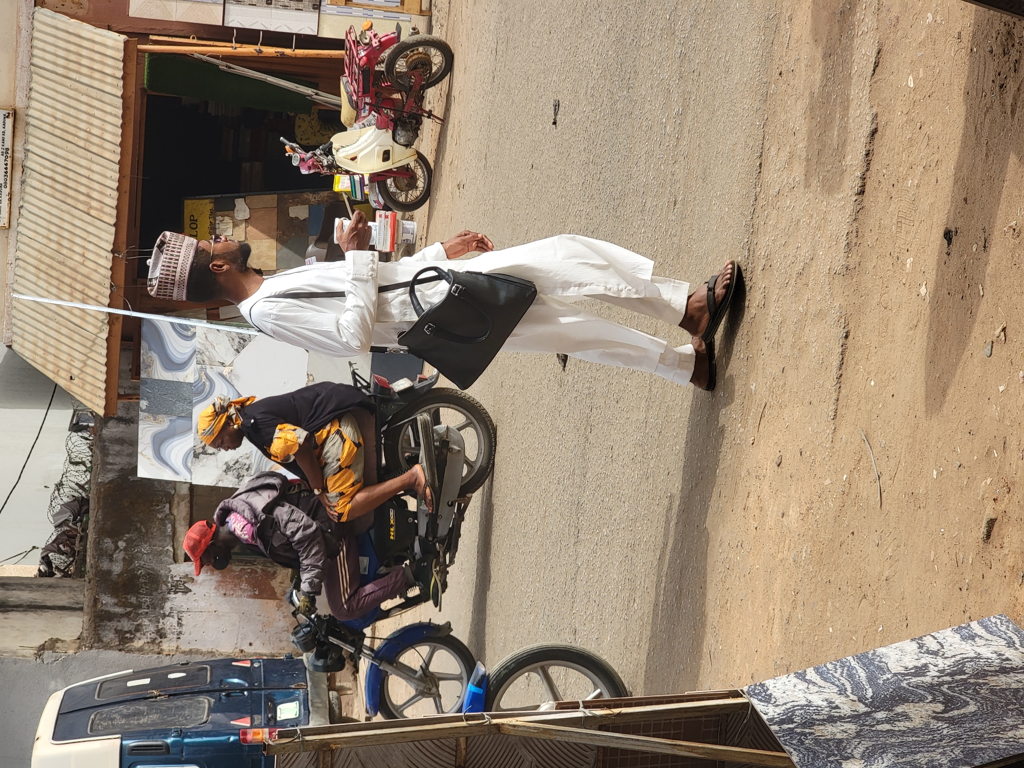 A street scene in Maiburiji, Nigeria, shows a man in white traditional attire with a black bag looking upwards. A man and a woman are on a motorcycle behind him. A van, tiles, and motorcycles are also visible.