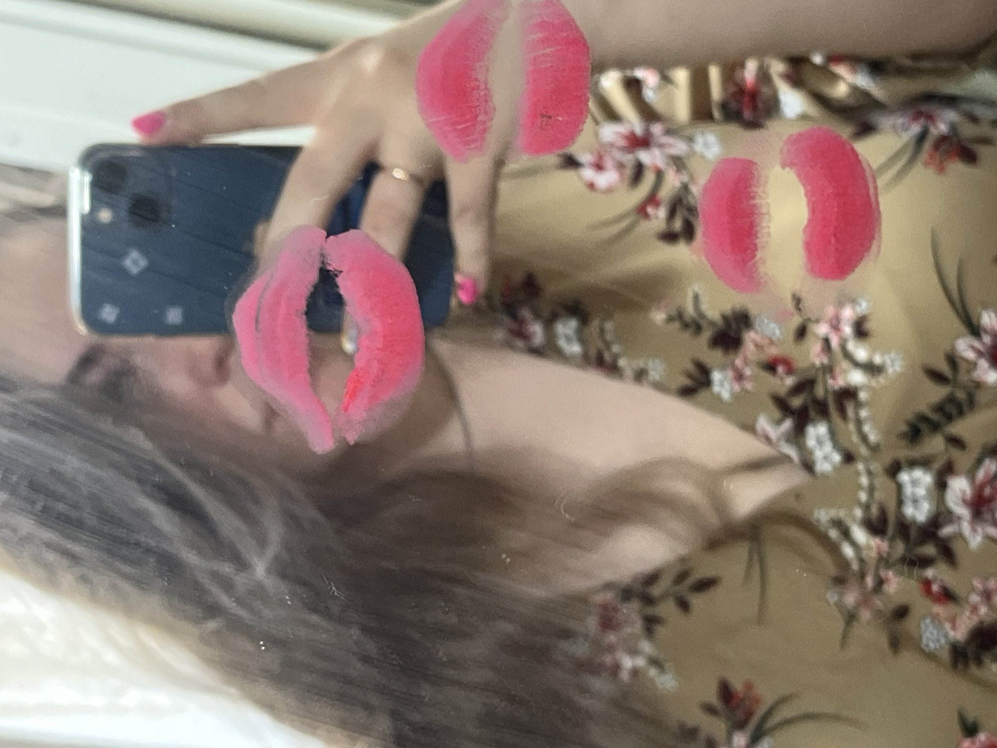 An angled mirror selfie captures a person reflected, holding a dark-colored smartphone, likely an iPhone, in their left hand to take the picture. The person has long, dark hair flowing around their face. Their visible hand sports bright pink nail polish and a simple ring on one finger. Three distinct, bright pink lipstick kiss marks are smeared across the mirror's surface: one prominently near the center-left, one in the upper-middle, and another mid-right. The reflection also shows the person wearing clothing with a floral pattern featuring pink, white, and dark red flowers with green leaves on a light brown or beige background. The setting appears to be indoors. There is no visible text in the image.