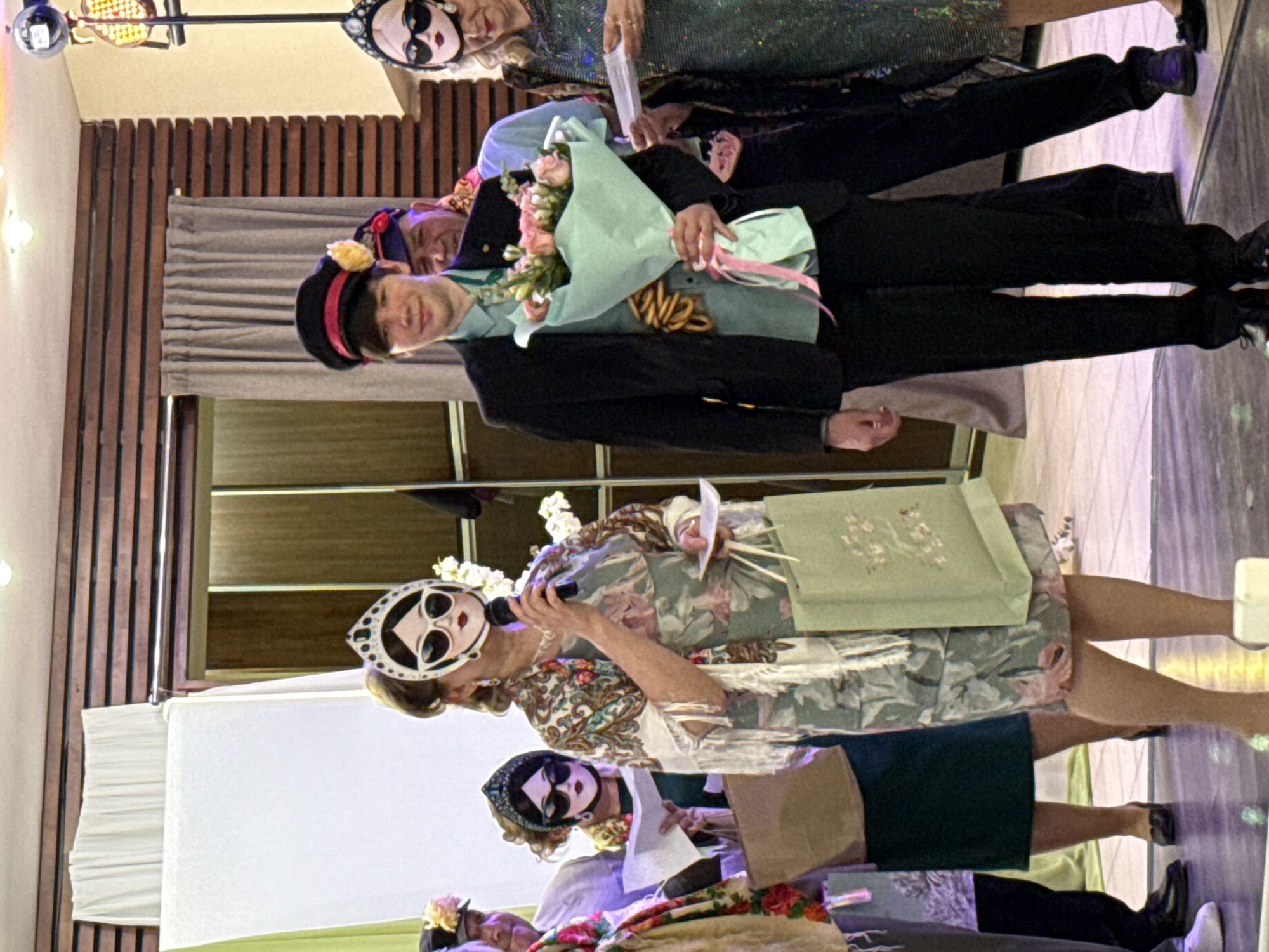 This image, captured at an indoor event in Chaikovsky, Russia, depicts a festive gathering, possibly an awards ceremony or themed performance. The photo is presented with a vertical orientation, but the content shows subjects standing upright.
Central to the frame is an individual, likely a young man, dressed in a dark suit jacket, a light blue collared shirt, and dark pants. He holds a large bouquet of pink roses wrapped in light blue paper and wears a dark hat with a yellow adornment. A sash crosses his chest, displaying unclear text that appears to be "W8S" or a similar combination of letters and numbers. He smiles pleasantly at the viewer. Behind him, another person with a dark hat and red band is visible, also smiling.
To the right of the central figure, a woman in a floral-patterned dress holds a microphone and a piece of paper, appearing to speak or host. She wears a distinctive white headpiece featuring a stylized face mask with large black sunglasses and red lips. A light green gift bag or box rests beside her. Further back, another woman is seen wearing a similar black stylized mask, holding papers. Partially visible in the top part of the image is another person wearing a white stylized mask and a sparkly dark green top.
The setting includes wooden wall paneling, a gray curtain, and what appears to be a stage or raised platform. The floor is somewhat reflective, and decorative lights are visible overhead. The overall ambiance is celebratory, with participants displaying positive emotions. The stylized masks and elaborate hats suggest a distinct, possibly retro or whimsical theme for the event. The time of day is indeterminate, but indoor lighting suggests it is likely evening.