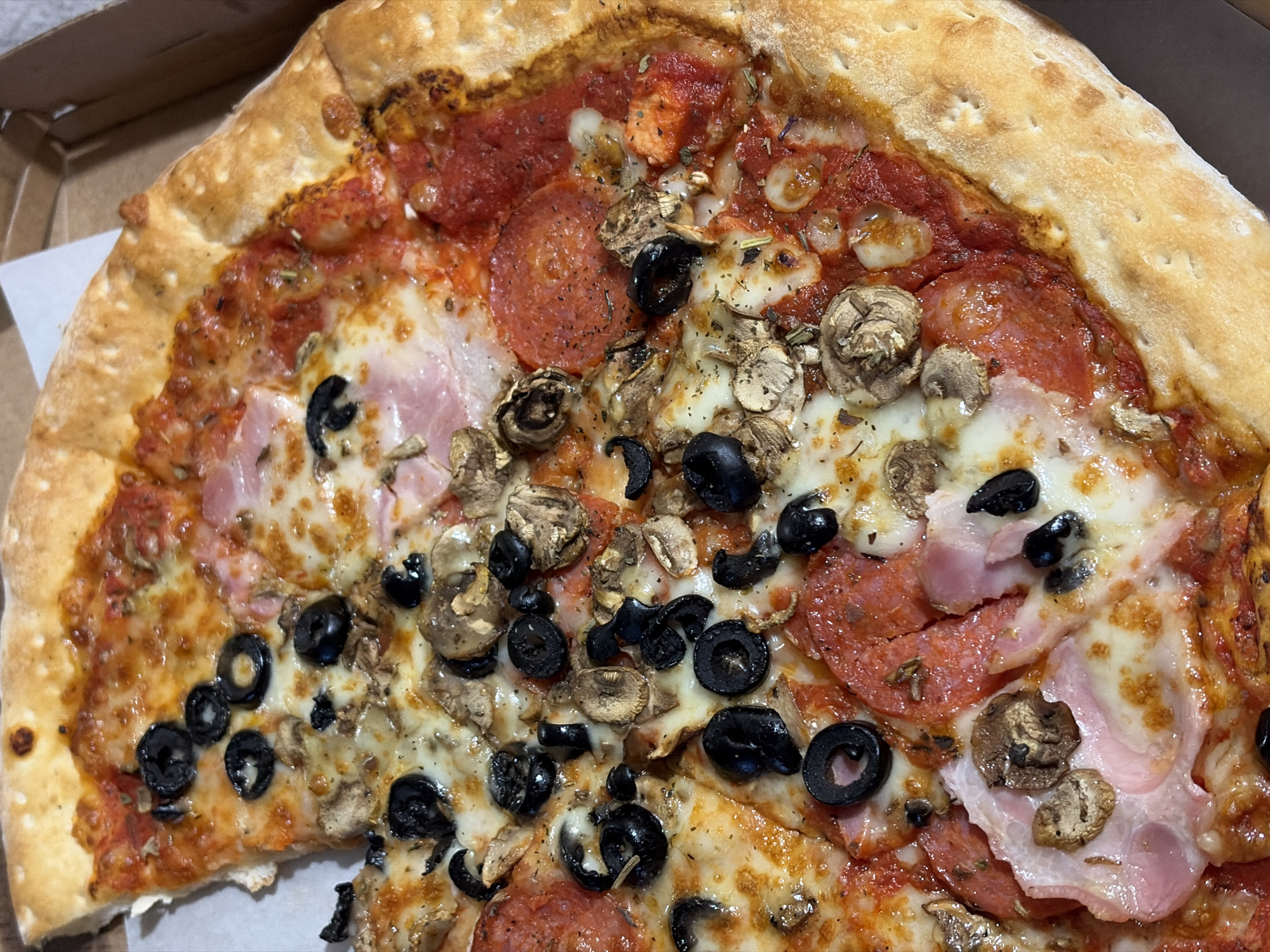 A close-up shot reveals a partially eaten pizza in a cardboard box. The pizza features a thick, golden crust and is topped with tomato sauce, melted cheese, pepperoni, ham, sliced black olives, and sliced mushrooms. The location is not specified.