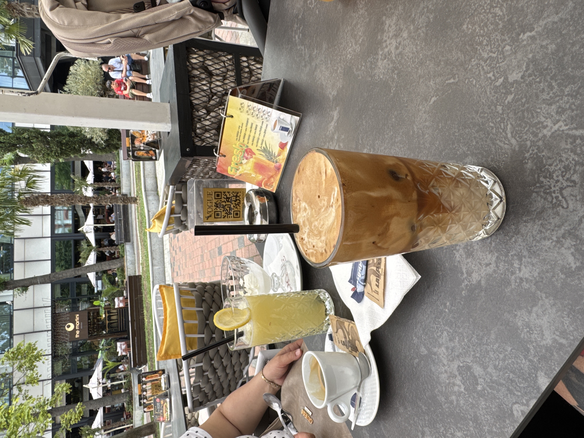 The image shows an outdoor seating area at "The Martini" cafe in Varna, Bulgaria. A table in the foreground displays several drinks and snacks: a tall glass of iced coffee with foam, a glass of yellow juice with a lemon slice and straw, a small cup of espresso with a saucer and spoon, and a small bowl of ice cream. Several sugar packets and napkins are also on the table. In the background, there is a building with signage for "The Martini," palm trees, umbrellas, and people seated at tables. A person's arm and hand are visible on the left side of the image, resting on the table. Another person, a man with a child, is sitting on a bench in the background, looking at their phones. The environment appears to be a sunny day in a commercial or recreational district.