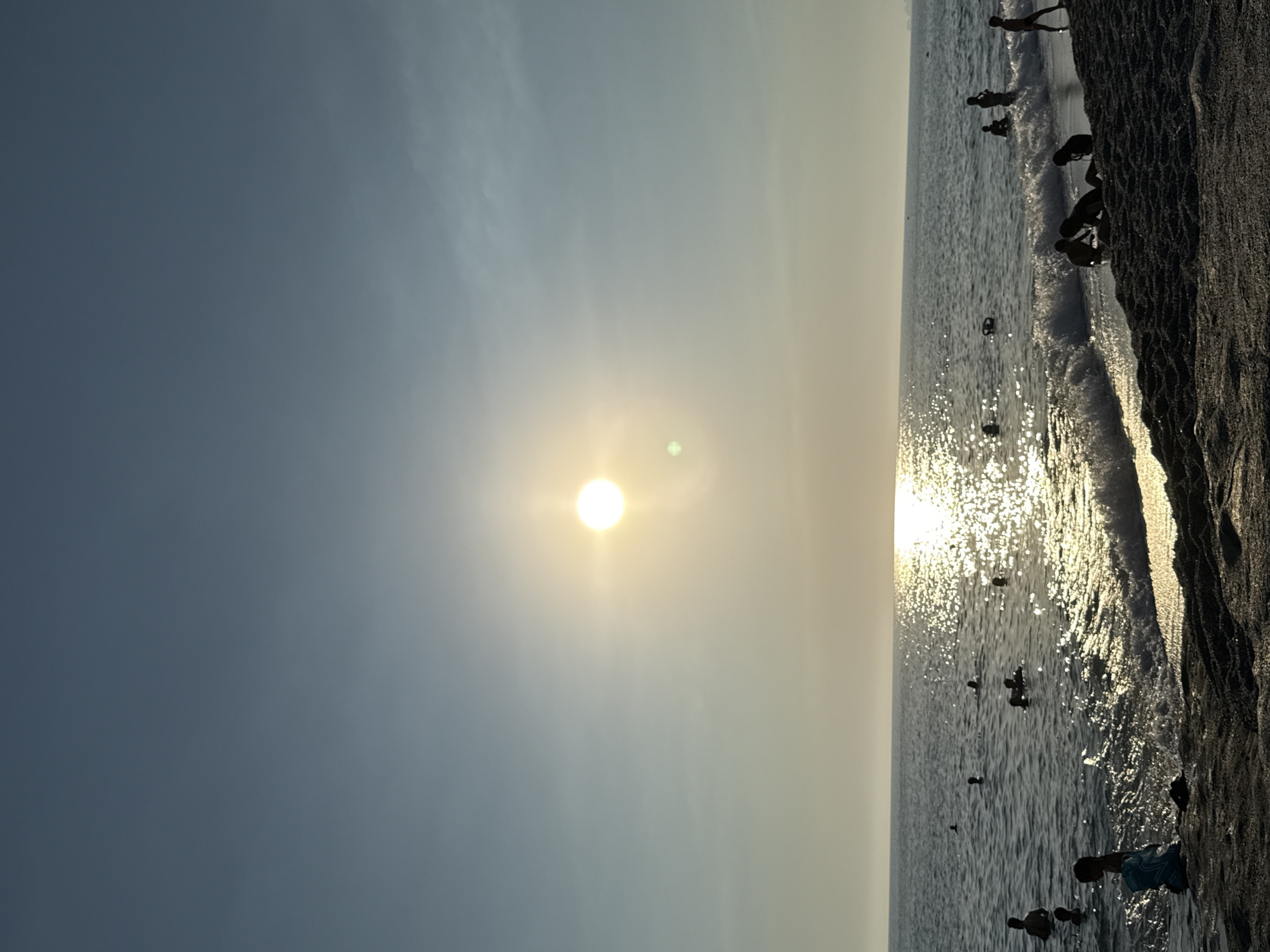 The image depicts a beach scene with people swimming and playing in the sea during daylight. The sun is visible in the sky, casting a bright reflection on the water. The foreground shows a sandy beach with some dark sand and shadows. The sea is calm with small waves breaking on the shore. Several individuals, appearing as silhouettes against the bright sun and water, are scattered throughout the water. One person is standing on the beach in the lower left corner, facing away from the camera, with a blue bag at their feet. Another group of people are visible near the shore on the right side of the image. The overall scene suggests a recreational setting at a beach, likely in Alanya, Turkey, given the bright sun, clear sky, and the presence of people enjoying the water.