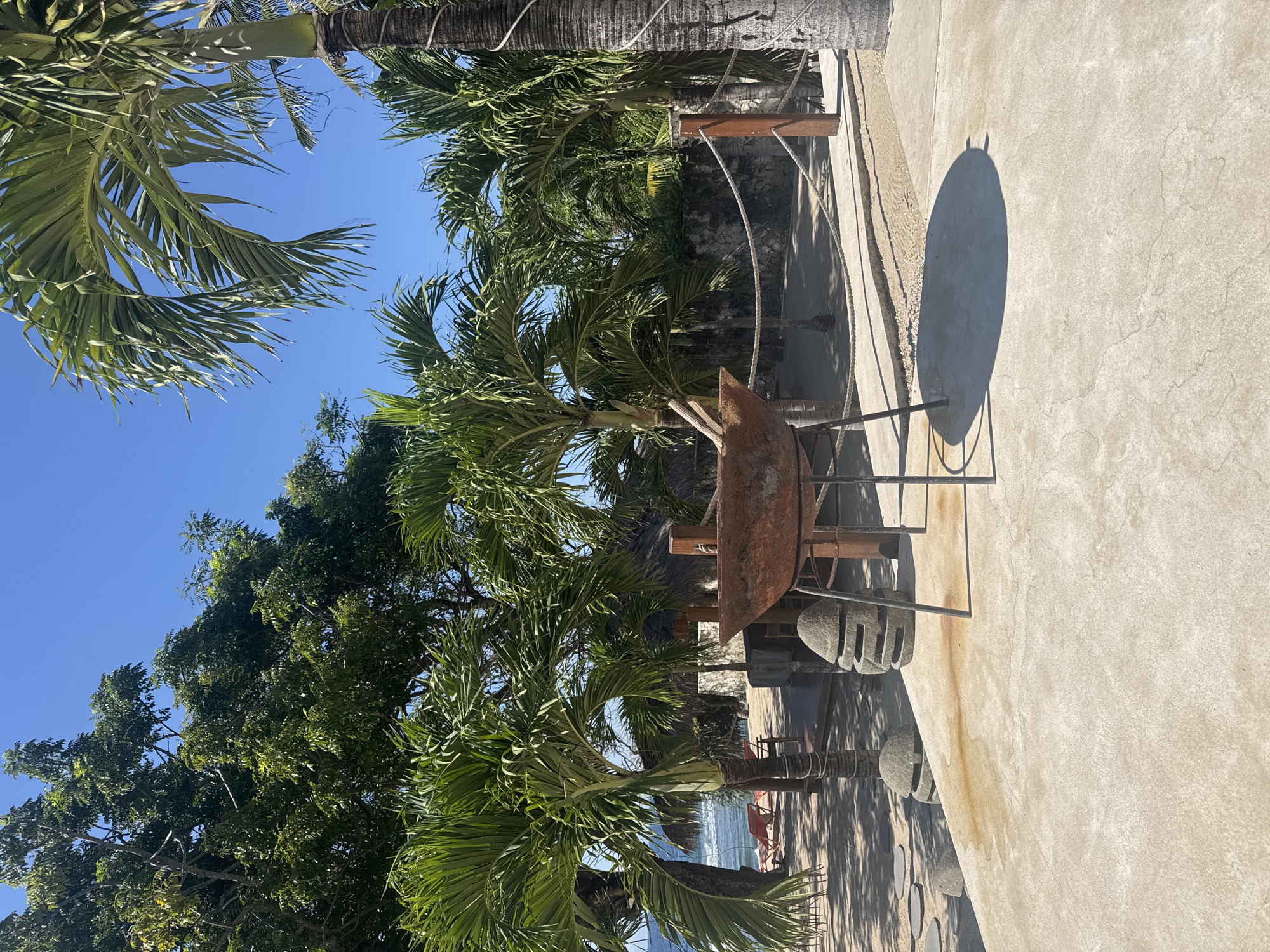 This image displays an outdoor tropical environment in Nusa Penida, Indonesia, under a clear, bright blue sky. The foreground is dominated by a light-colored concrete patio, exhibiting minor cracks and discolored patches. A large, rusty, bowl-shaped metal object, supported by three black metal legs, is centrally positioned on this patio, casting a distinct elliptical shadow to its right. To the left of this object, on the patio's edge, stand two grey, stacked stone decorative elements with horizontal apertures.
Beyond the concrete patio, a sandy area is visible, interspersed with several dark, circular stepping stones. This area is densely populated with various green tropical trees, including numerous palm trees with textured trunks and prominent fronds. Through a gap in the foliage on the far left, a section of the ocean with visible waves and two red beach chairs on the sand is discernible. Further into the midground, several structures featuring light-colored walls and dark thatched roofs are situated amidst the trees. A rope barrier, secured by dark wooden posts, extends horizontally across the sandy ground. No individuals are present in the scene. The entire area is illuminated by strong natural sunlight, producing sharp shadows.