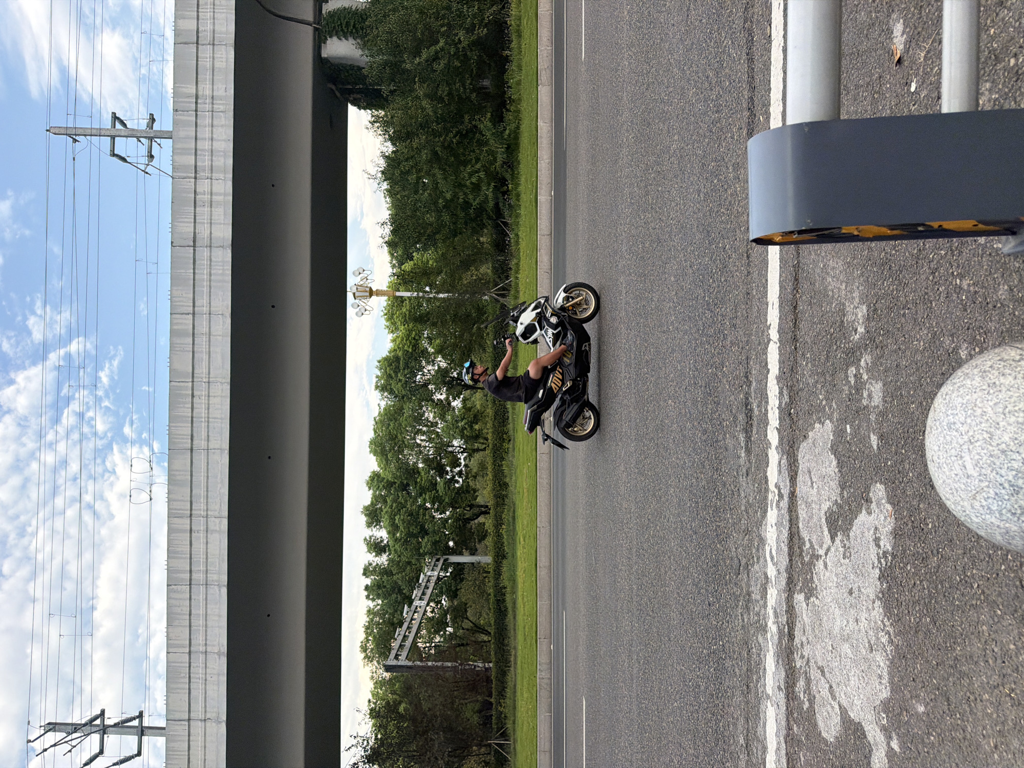 The image depicts a man riding a scooter on an asphalt road under a large overpass. The overpass is a concrete structure with visible power lines above it. The road is bordered by a grassy median and trees. The scooter is black and white with gold accents, and the rider is wearing a helmet and shorts. The sky is partly cloudy. The foreground shows a portion of a white line on the road and a grey circular object. This scene is located on a road in Jiangjin, China.