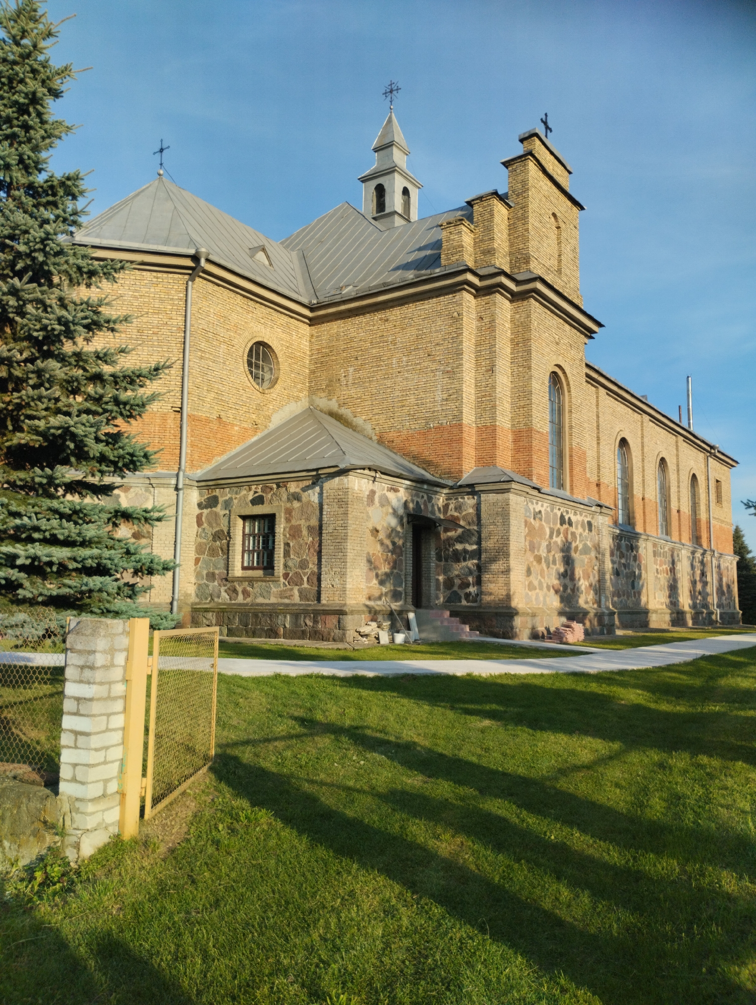 This is a full-shot, eye-level, outdoor photograph of a brick church with a grey metal roof. The church has a large, prominent tower with a cross at the apex and a smaller bell tower. The main facade is made of light-colored bricks with a decorative band of darker bricks partway up. The lower section of the church and the entrance are constructed with large, irregular stones. A large evergreen tree is visible on the left side of the frame, partially obscuring the church. In the foreground, a green lawn is present, with a concrete path leading around the church. A fence with yellow posts and wire mesh is also visible on the left. The sky is clear and blue. The scene appears to be set in a rural area, likely the village of Vishnevo (Вішнеўскі сельскі Савет), Belarus, based on the surrounding architecture and vegetation. There are no people or animals visible in the image. The lighting suggests it is late afternoon, with long shadows cast across the lawn.