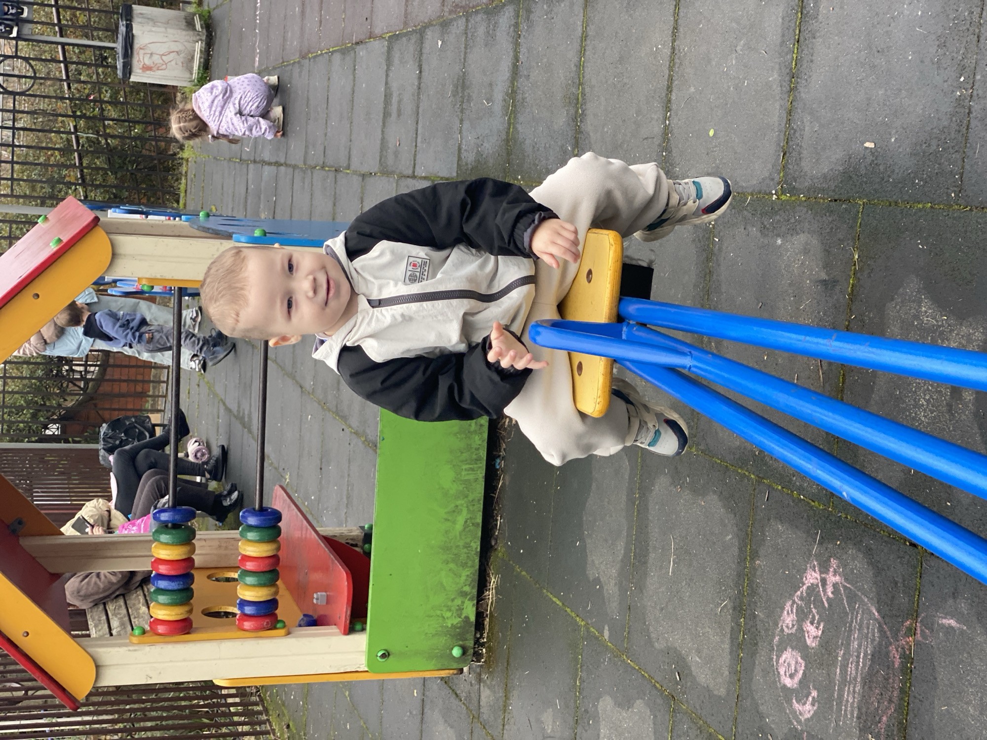 A young boy in a black and white jacket and beige pants is sitting on a blue and yellow seesaw in a playground. He is facing the camera and smiling. Behind him is a play structure with colored rings. In the background, other people are sitting on a bench, and a child in a purple outfit is crouching on the ground. The playground is located in Sochi, Russia, as evidenced by the context of the image. The ground is paved and there is a chalk drawing. A metal fence and vegetation can be seen in the background.