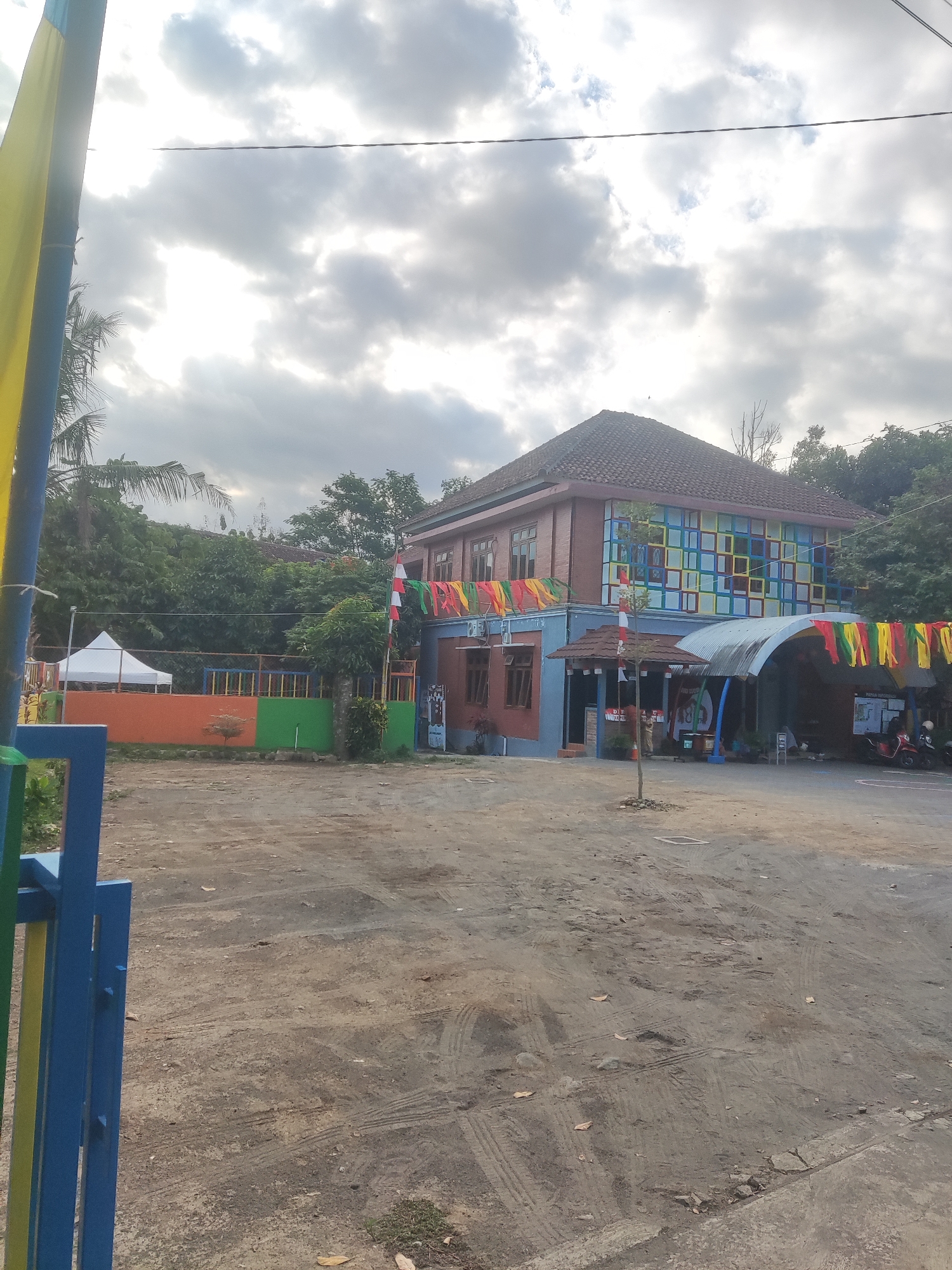 The image depicts a multi-story building with a red tile roof, likely a school or community center, in Trihanggo, Indonesia. The building's facade features brightly colored square patterns, with blue, yellow, and red dominant. Festive streamers in yellow, red, and green hang across the front of the building, and a small white tent is visible to the left. The ground in front of the building is a dirt or gravel lot with tire tracks and scattered leaves. Trees and foliage surround the structure. A blue and yellow fence is prominent in the foreground, suggesting it is an entrance to the property. Two motorcycles are parked under an arched, corrugated metal awning to the right of the building. The sky is overcast with diffused sunlight.
