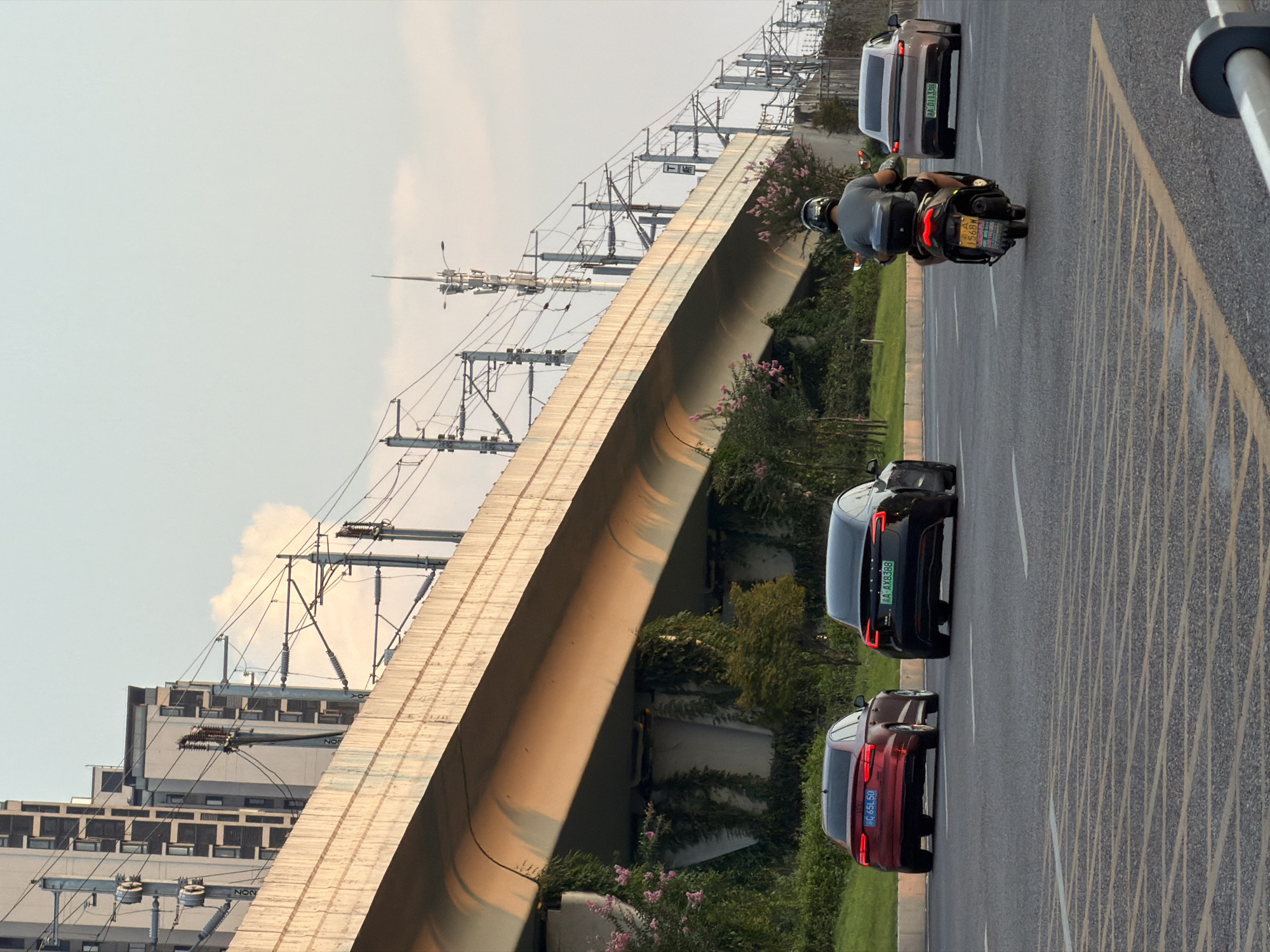 A wide-angle, eye-level shot captures a street scene in Jiangjin, China, with an elevated highway or bridge dominating the upper portion of the frame. The sky is a pale blue with a hint of clouds. Below the elevated structure, several vehicles are in motion on a multi-lane asphalt road.
On the left, a red sedan with license plate "C 65L50" is driving forward. Next to it, a black sedan, possibly a Tesla, with license plate "A AXB388" is also proceeding forward. In the center of the image, a person wearing a gray shirt and a helmet rides a scooter with a black storage box on the back. The scooter's license plate is "A 156BW". To the right, a light-colored sedan with license plate "A 011336" is visible, also driving forward.
The elevated structure is supported by concrete pillars, some of which are covered in green ivy and pink flowering plants. Electrical pylons and overhead wires associated with a railway or power lines run along the length of the elevated structure. In the background, modern high-rise buildings are visible. The road has yellow and white lane markings, with a distinct pattern of yellow diagonal lines in the foreground. The lighting suggests it is late afternoon or early evening.
