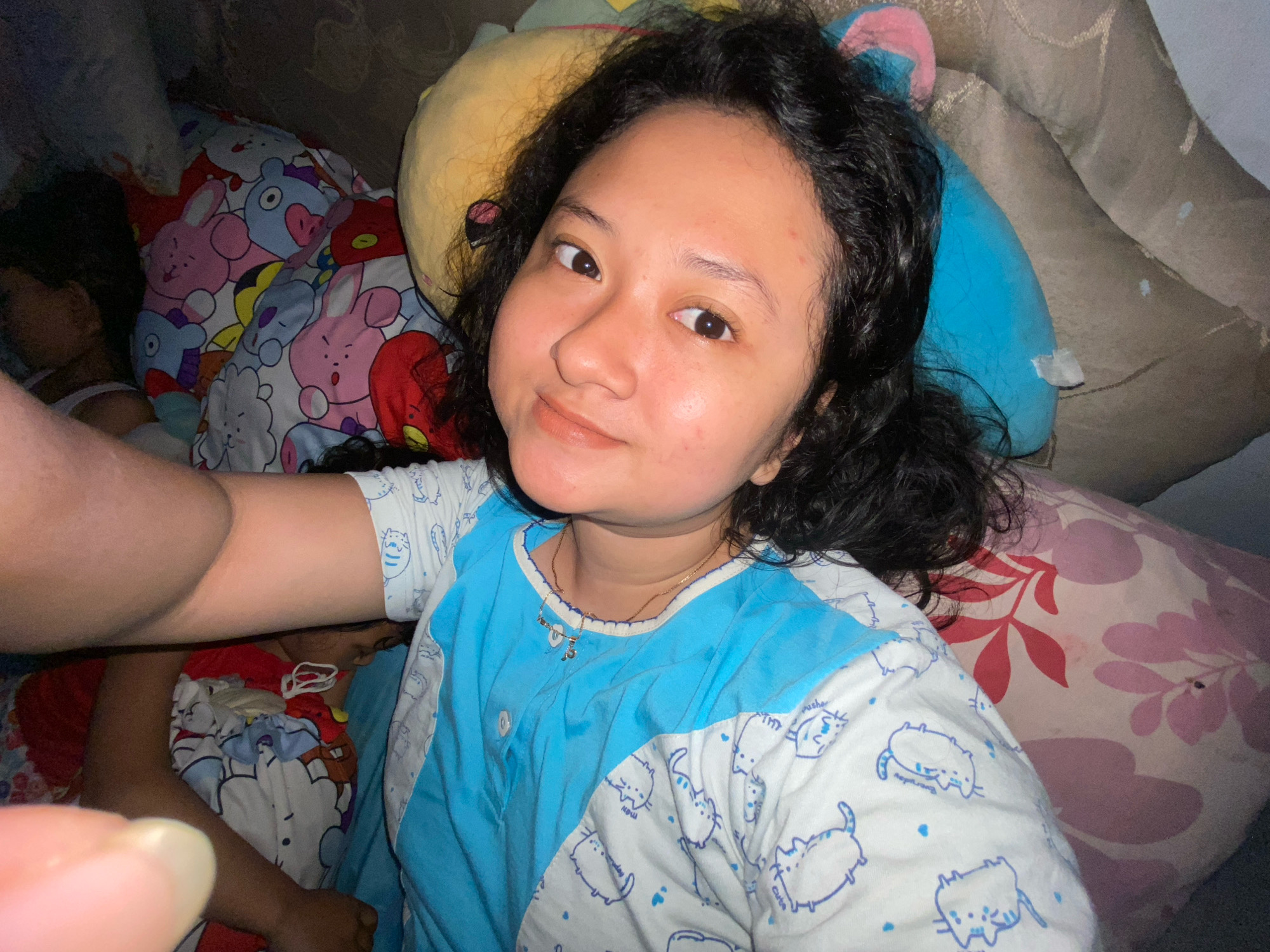 A young Indonesian woman with curly black hair is lying on a bed, surrounded by stuffed animals. She is wearing a blue and white pajama top adorned with cartoon cat prints. Her face is turned towards the camera, and she has a gentle smile. The background is filled with various plush toys, including one with a yellow face and another with blue ears. Part of a person's arm and hand are visible in the lower left of the frame, and another person is lying down in the background, partially obscured by stuffed animals. The scene suggests a relaxed, indoor setting, possibly a bedroom. The location is identified as Gelam, Indonesia.