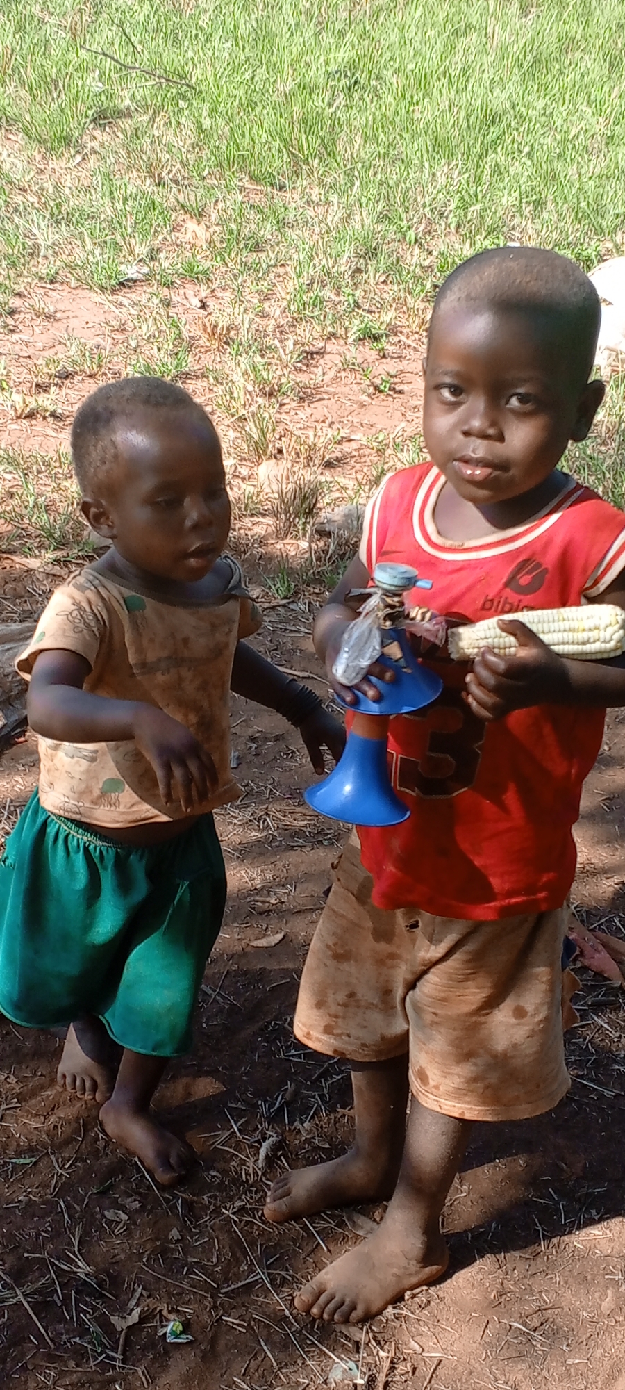Two young children stand outdoors in Ngalula, Democratic Republic of the Congo. The child on the left wears a yellowish-brown t-shirt and green shorts. The child on the right wears a red t-shirt and brown shorts. The latter is holding a blue plastic toy in one hand and an ear of corn in the other. The ground is a mix of dirt and sparse vegetation.