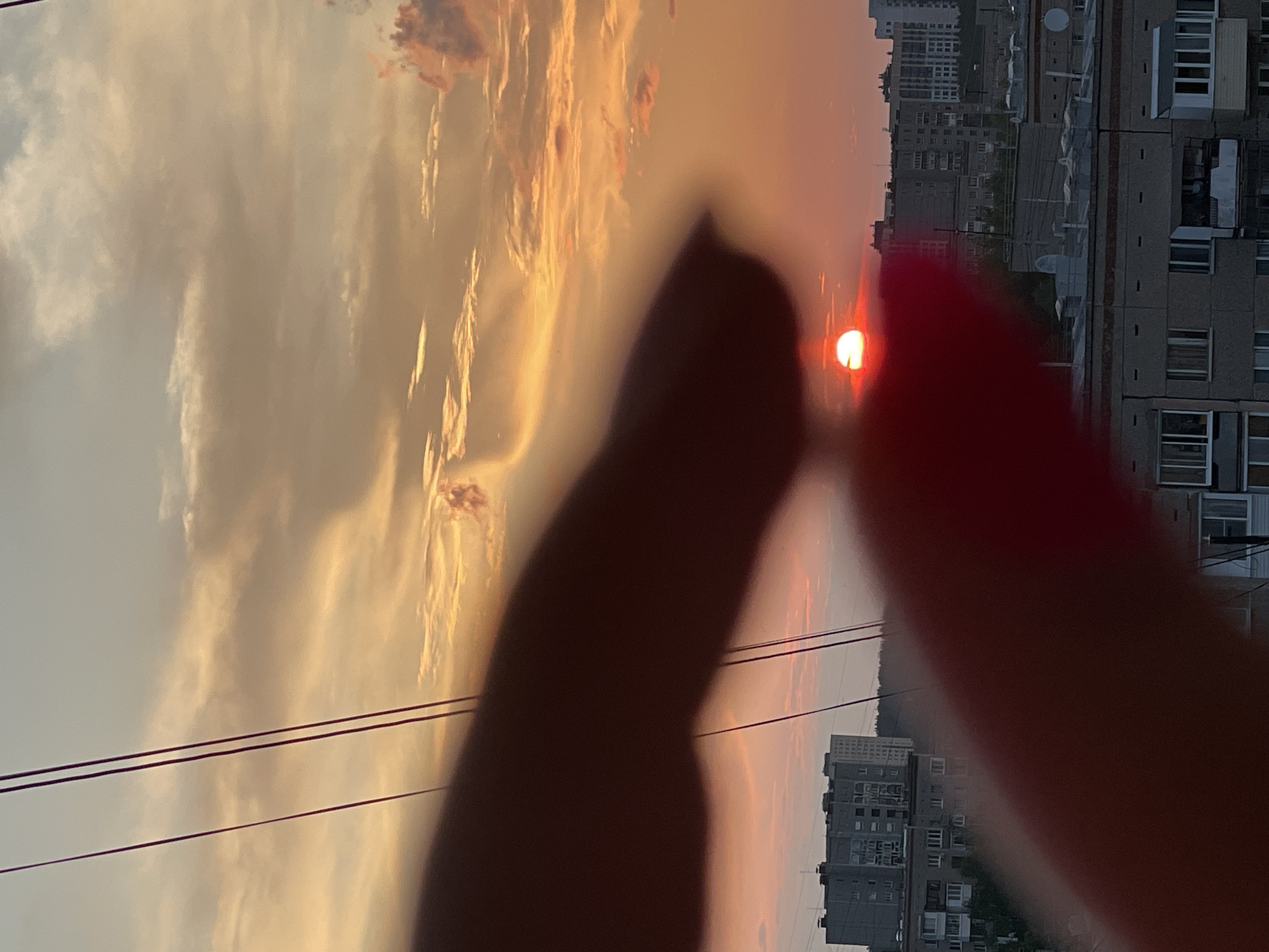 This image captures a sunset over the city of Izhevsk, Russia. The foreground is dominated by the blurred shapes of fingers, one dark and one with red nail polish. The sun, a bright orb of red and yellow, is partially obscured by clouds and is setting behind a cluster of residential buildings. The sky is filled with dramatic, colorful clouds ranging from pale yellow to vibrant orange and pink. Several power lines crisscross the sky. The buildings in the background appear to be typical Soviet-era apartment blocks, some with visible balconies and windows. The overall impression is a personal, candid moment framed against a striking natural phenomenon.