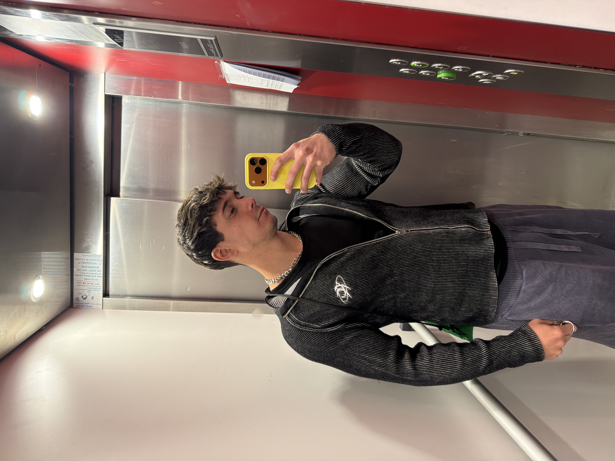 A young man is taking a mirror selfie inside an elevator, located in Sofia, Bulgaria. He is seen from the chest up, with his head slightly tilted as he looks at his bright yellow smartphone, held in his right hand. He has short, dark, curly hair and light stubble. He is wearing a dark, horizontally striped zip-up jacket with a white, circular logo (resembling the Vivienne Westwood orb) on the left chest, over a black top. A silver chain necklace is visible around his neck.
The elevator's interior features metallic walls and a red panel at the top. To the man's left, a circular light fixture is visible on the wall. A control panel with various buttons, including several green and white ones, is mounted on the red panel. Some white papers are tucked into the gap between the red panel and the metallic wall on the left.
Visible text on the papers, which is upside down and likely Cyrillic, includes fragments like "ИНТЕРНЕТ" (Internet), "ТЕЛЕФОН" (Telephone), and various numbers, as well as a partial web address ending in "pdn.bg," further supporting the Bulgarian context. The overall scene depicts a casual moment of self-photography within an indoor setting.