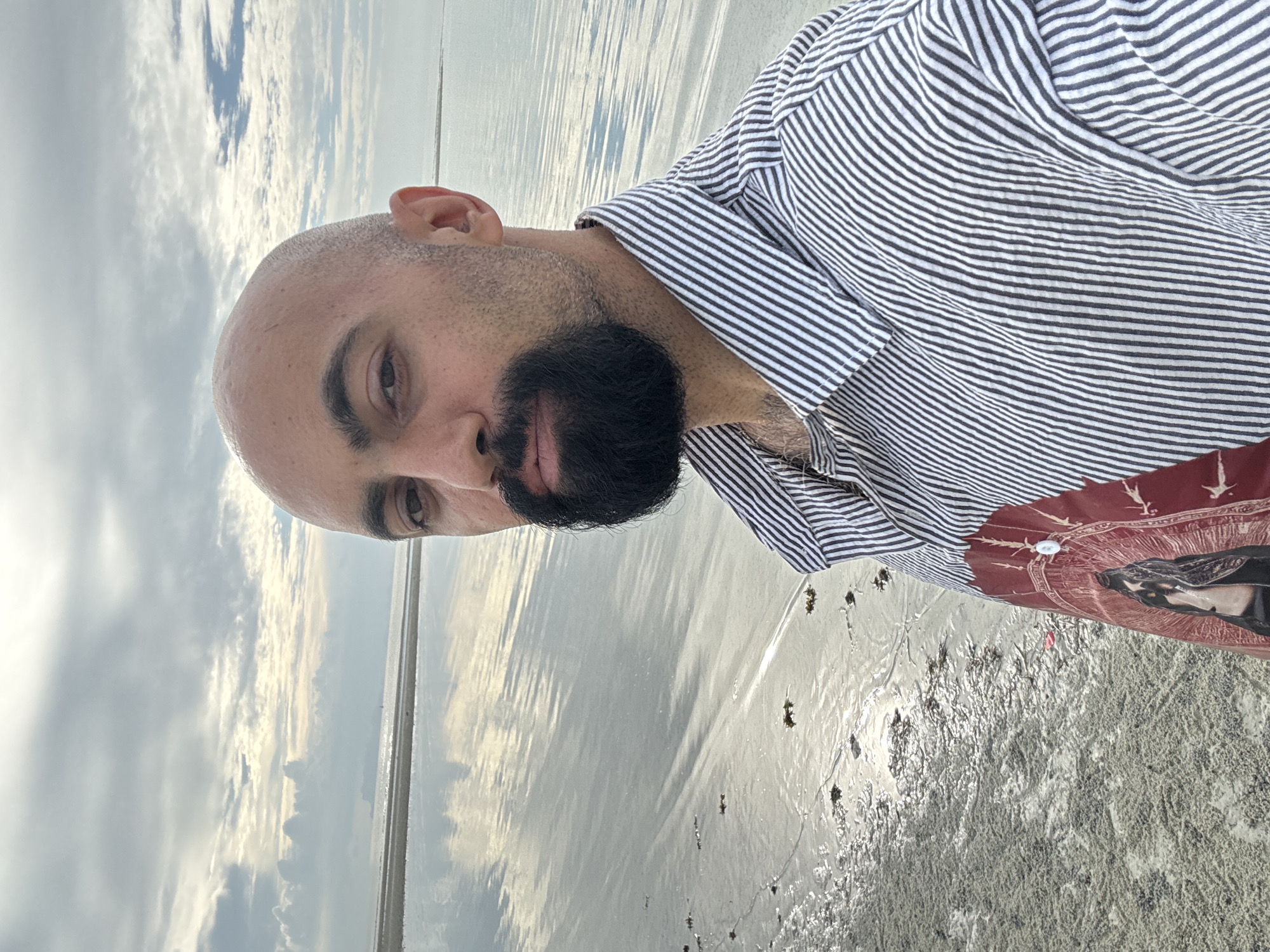 A bald man with a beard is taking a selfie on a sandy beach in Ko Samui District, Thailand. He is wearing a black and white striped button-down shirt with a graphic design on the front. The water is calm and reflects the cloudy sky. The sand is wet and has some seaweed scattered on it. In the distance, there is a faint outline of a landmass.