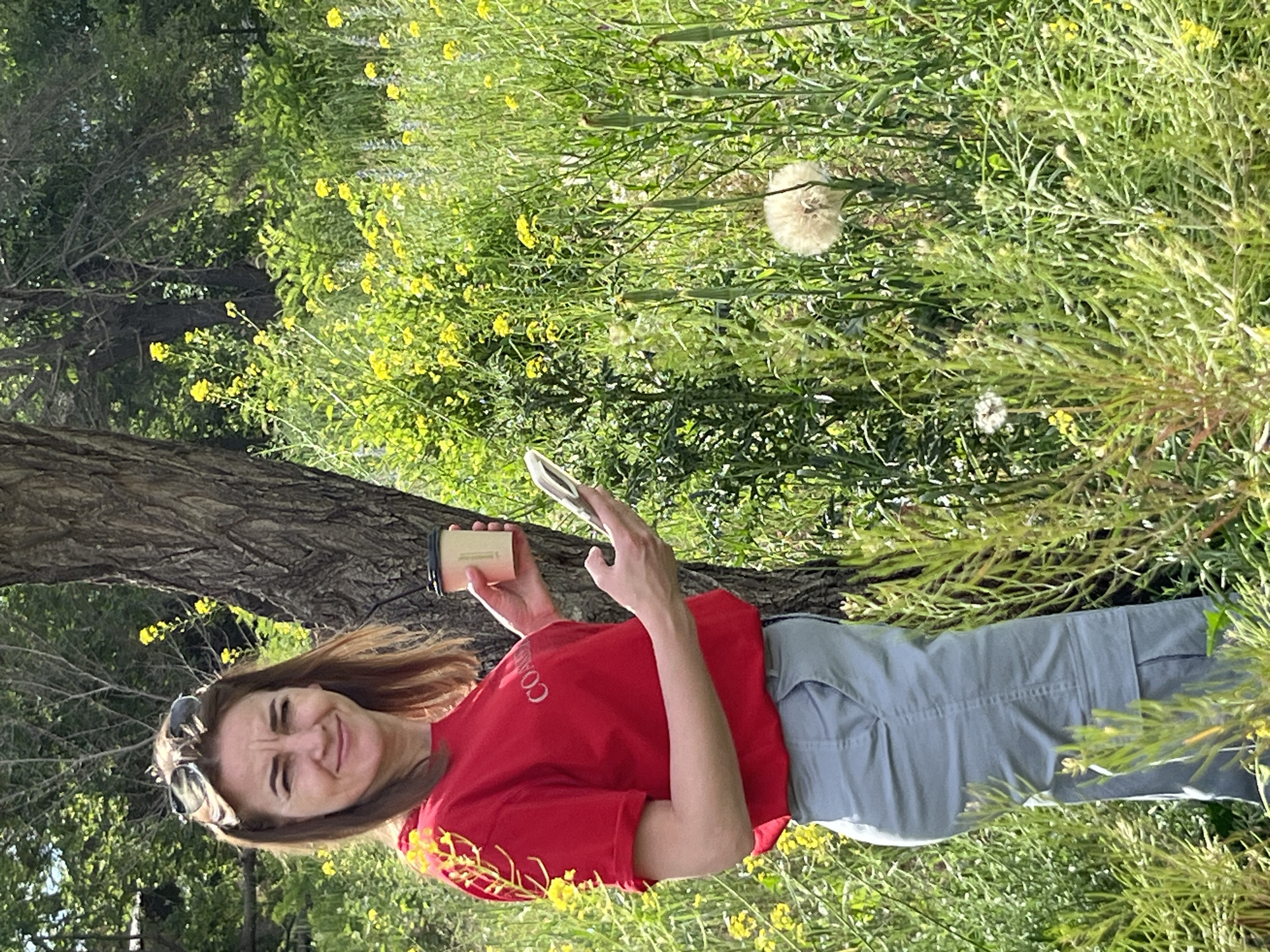 A woman stands in a grassy field with yellow flowers and a tree trunk to her right. She is wearing a red t-shirt and light blue cargo pants. She is holding a coffee cup in her right hand and a smartphone in her left hand. Sunglasses are perched on her head. The background consists of trees and more vegetation. The image is not specific to the city of Kryvyi Rih, Ukraine.