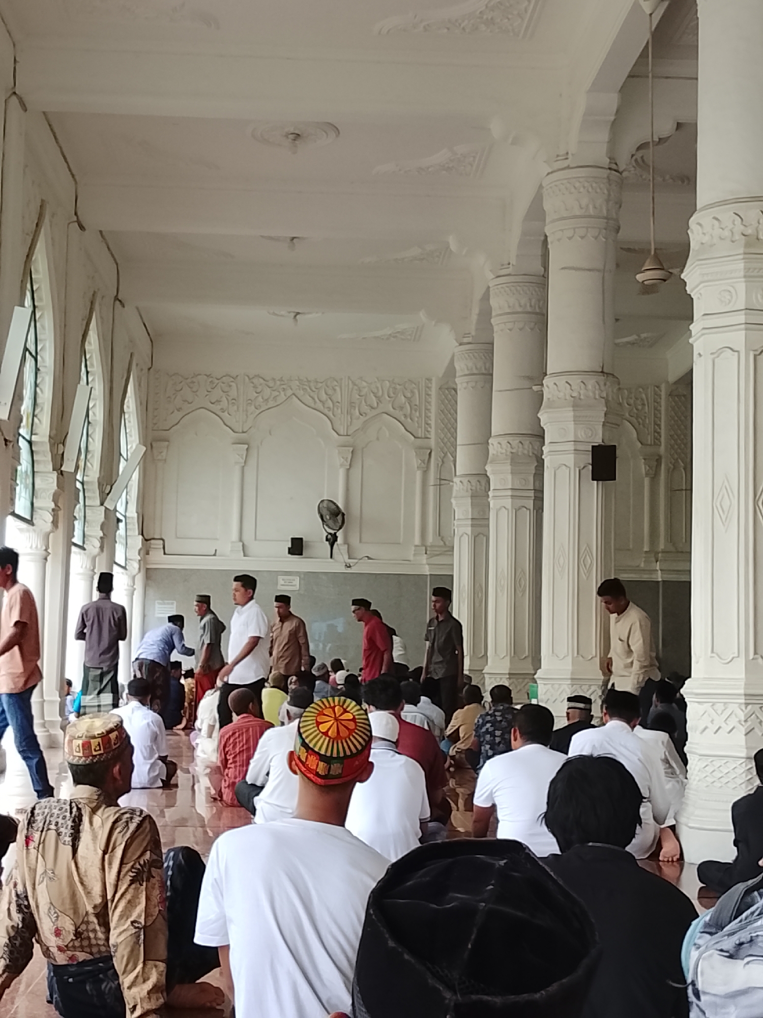 The image depicts the interior of a mosque in Lhokseumawe, Indonesia, likely during prayer or a religious gathering. A large group of men, dressed in traditional Muslim attire, are present. Many are seated on the floor, facing the same direction, indicating they are engaged in worship. The architecture features ornate white columns and decorative carvings on the walls and ceiling, suggesting a well-maintained and aesthetically significant structure. Natural light streams in from windows on the left, illuminating the space and highlighting the white surfaces. Some individuals are standing or walking, suggesting the event is ongoing or transitioning. The overall atmosphere appears peaceful and communal.