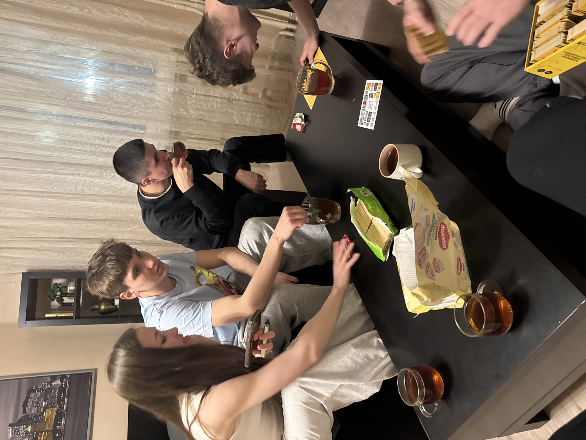 A group of approximately five young adults are gathered around a black rectangular table in a room. They are drinking tea from glass mugs and eating snacks, including waffles and cookies. One person is holding a smartphone and a vape pen. A game box is on the table. The room has beige walls, a dark gray shelving unit, and a framed picture of a city skyline. The location is inferred to be Kazan, Russia, based on the packaging labels of the food items.